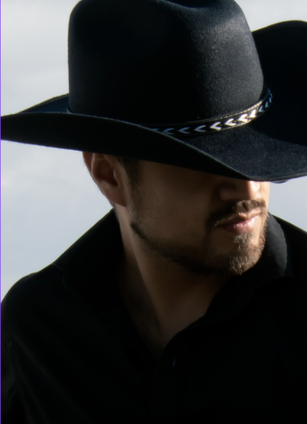 A man wearing a wide-brimmed black hat with decorative band, partially obscuring his face, with a light background.