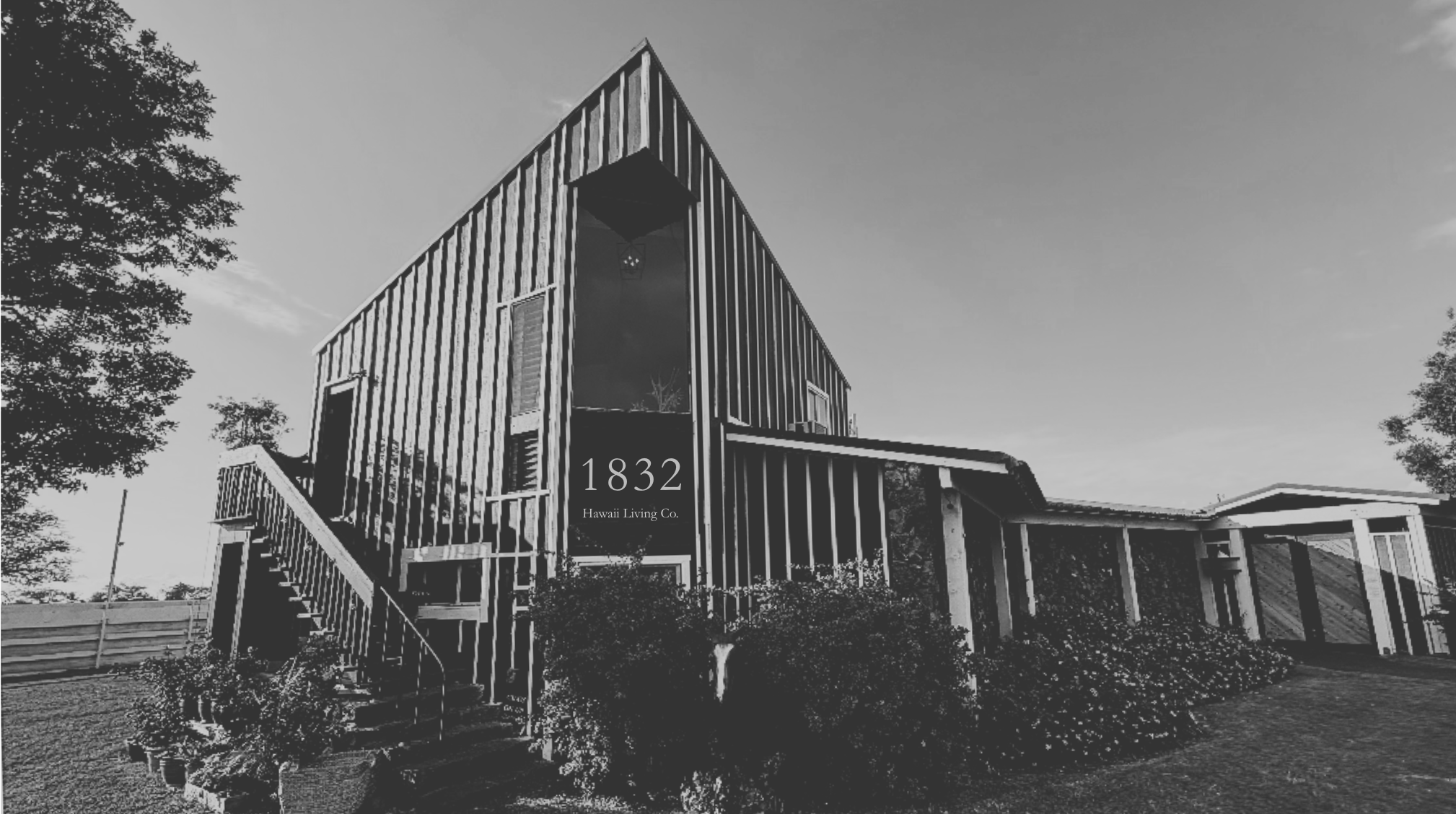 Black and white photo of a modern house with a tall, narrow front, labeled 1832 Hawaii Living Co., with stairs leading to the entrance and various windows, surrounded by trees and bushes.