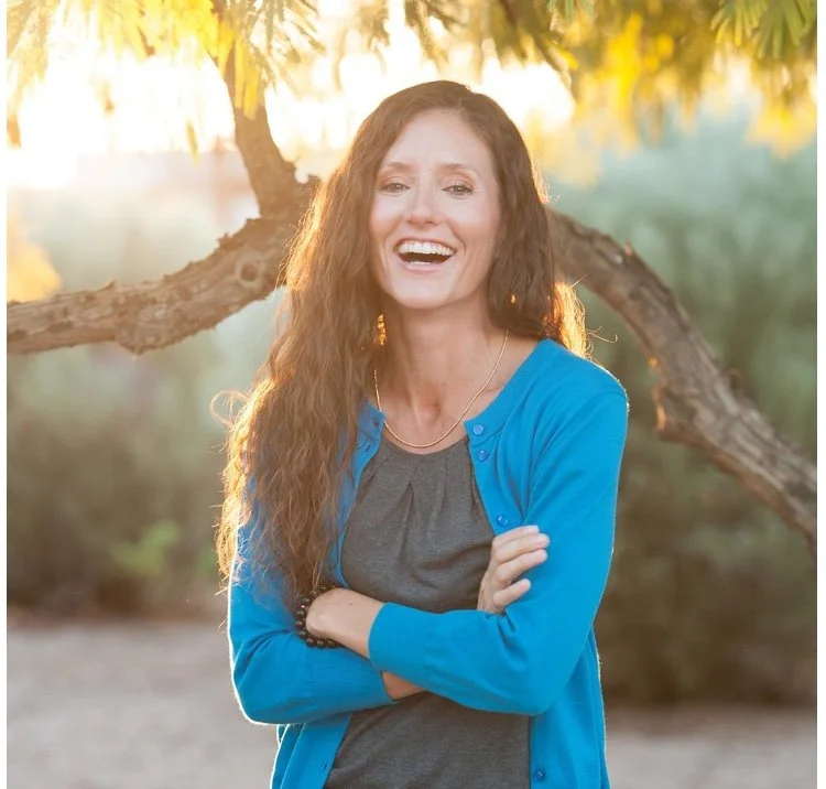 A woman with long, wavy brown hair smiling and standing outdoors under a tree at sunset, wearing a blue cardigan over a gray top.