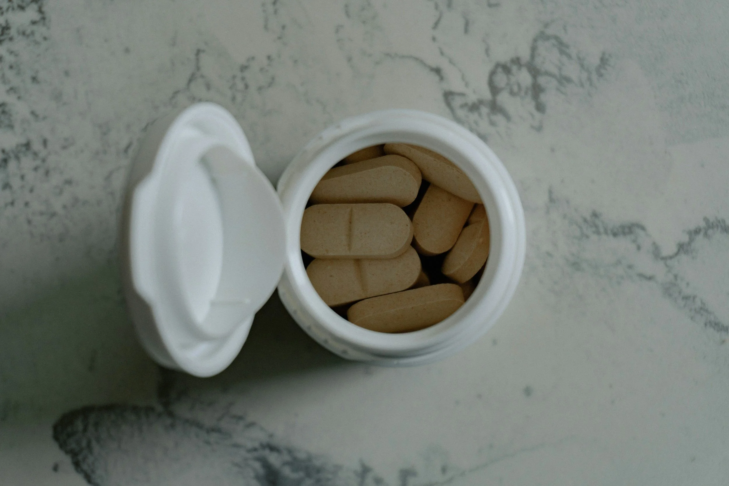 Open white pill bottle filled with beige tablets on a white surface.