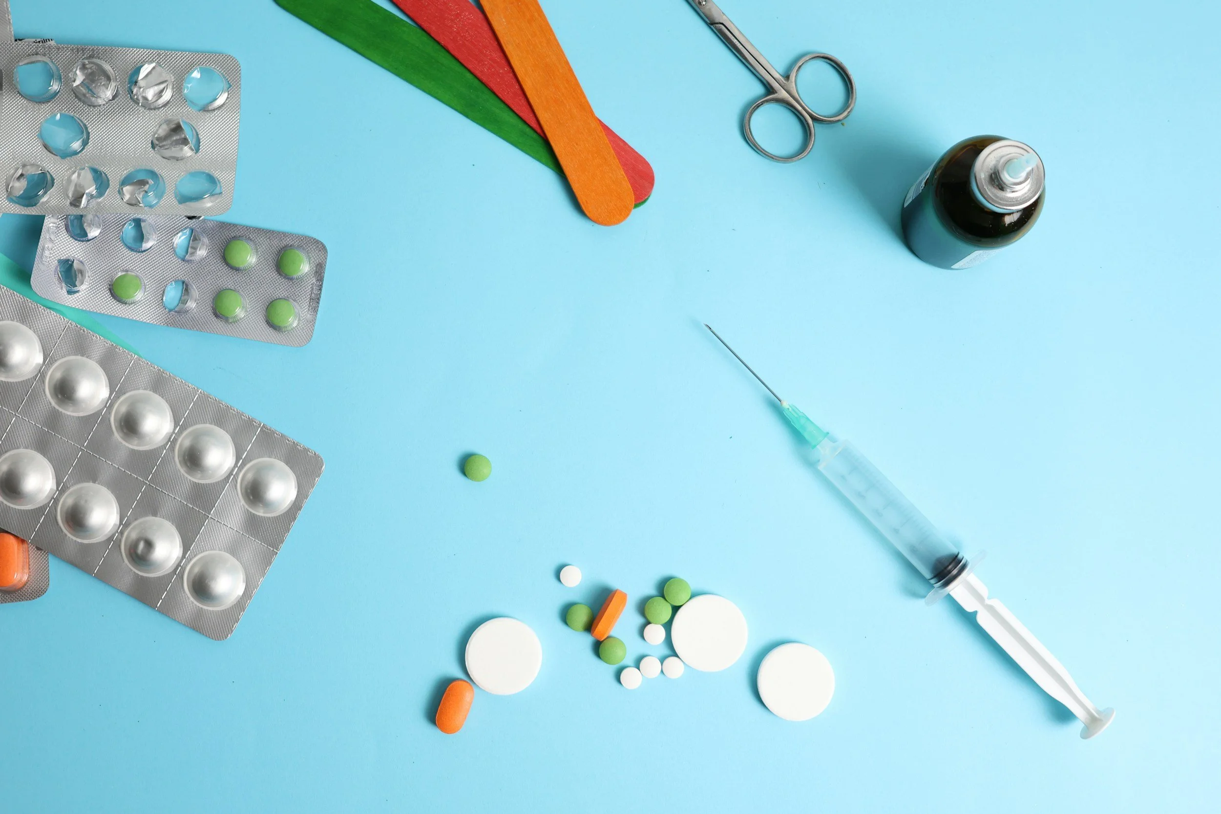 Medical supplies including blister packs of pills, loose pills, a syringe, a small dark bottle, scissors, and colorful wooden tongue depressors on a light blue background.