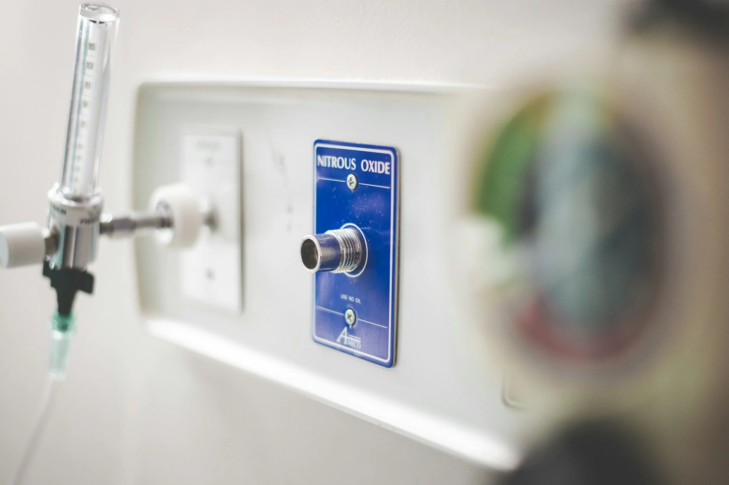 Close-up of a medical oxygen outlet with a blue label reading 'Nitrous Oxide' and a tube connected to the outlet, mounted on a white wall.