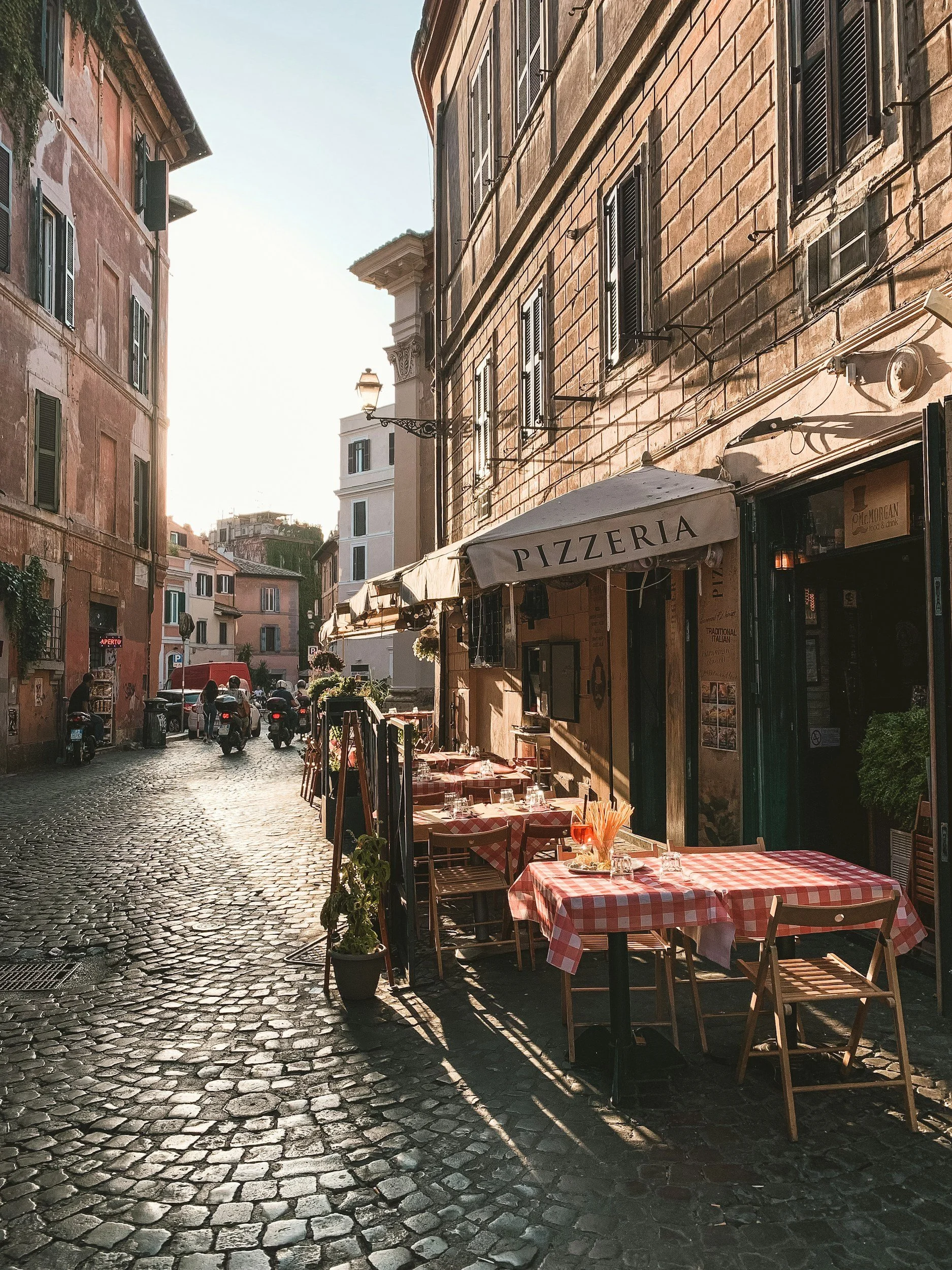 A narrow cobblestone street lined with restaurant tables covered with red and white checkered tablecloths, outside a pizzeria with outdoor seating. Buildings with shuttered windows and a streetlamp are visible in the background.