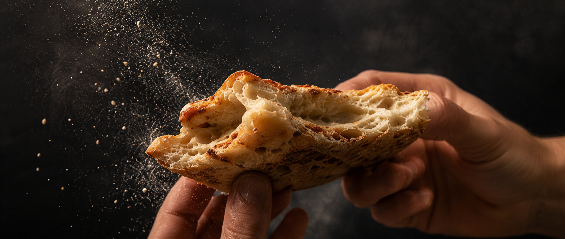 Person breaking open a piece of bread with flour falling out against a dark background.
