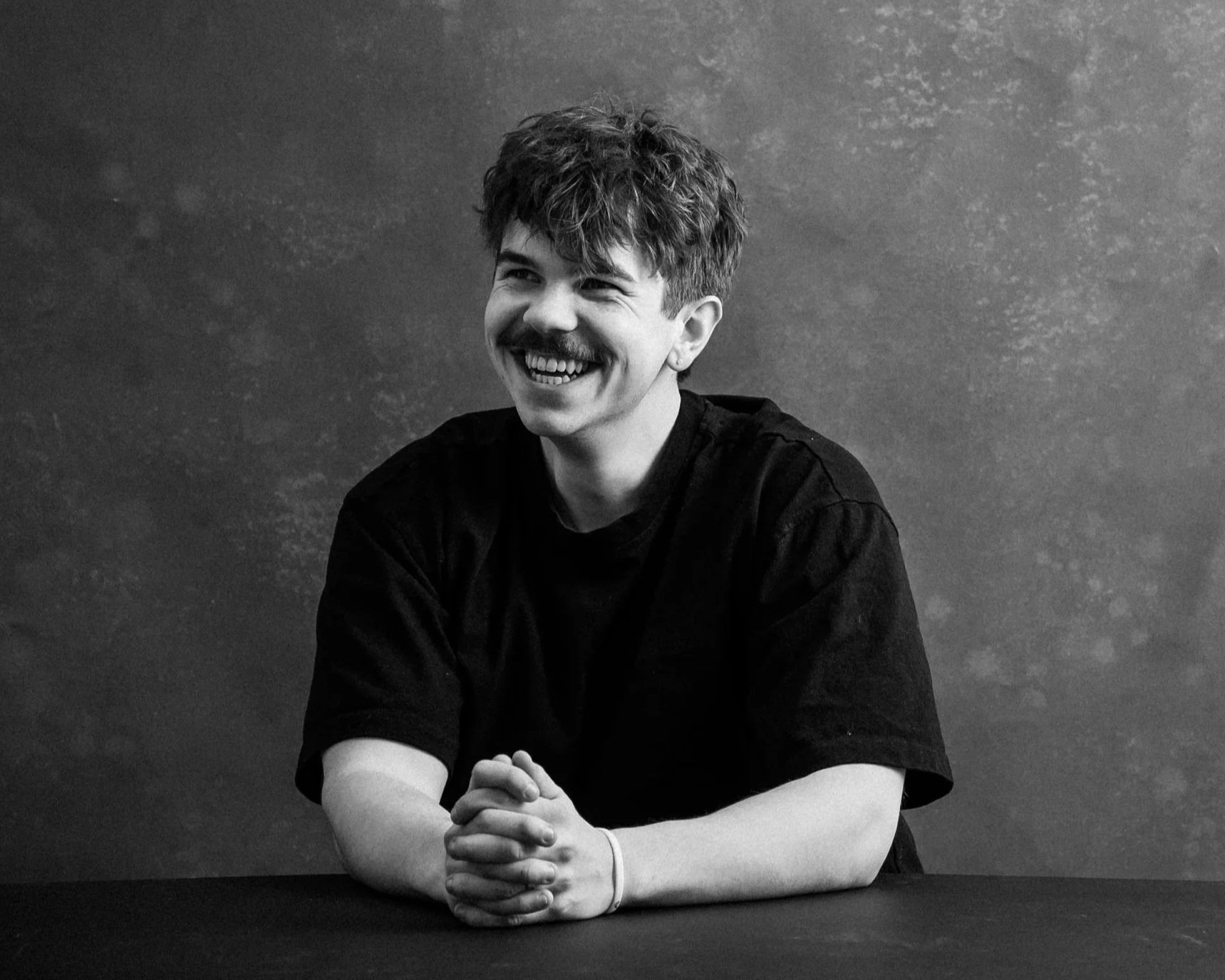Black and white photo of a young man smiling, sitting at a table with folded hands, against a textured dark background.