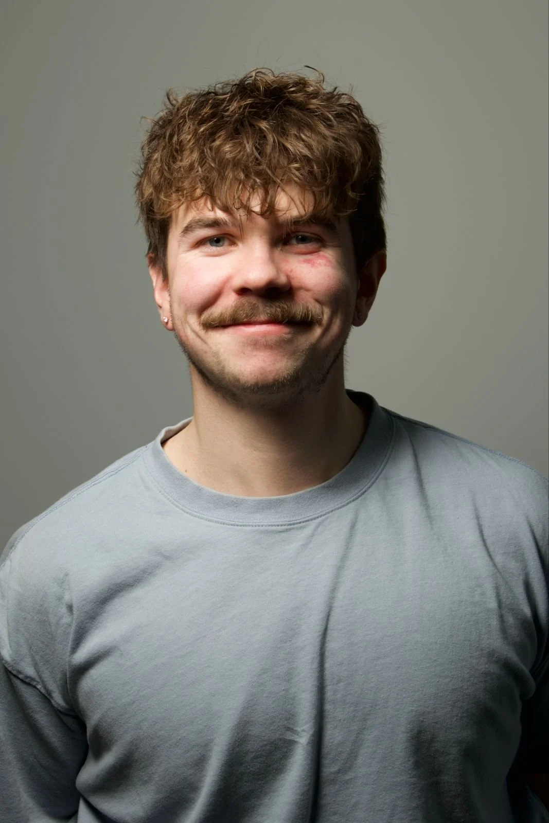 A young man with brown curly hair, and a moustache, smiling while wearing a light grey t-shirt against a plain background.