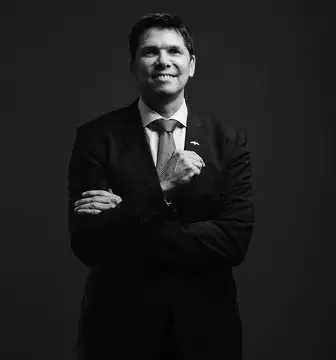 A man in a suit smiling and crossing his arms against a dark background.