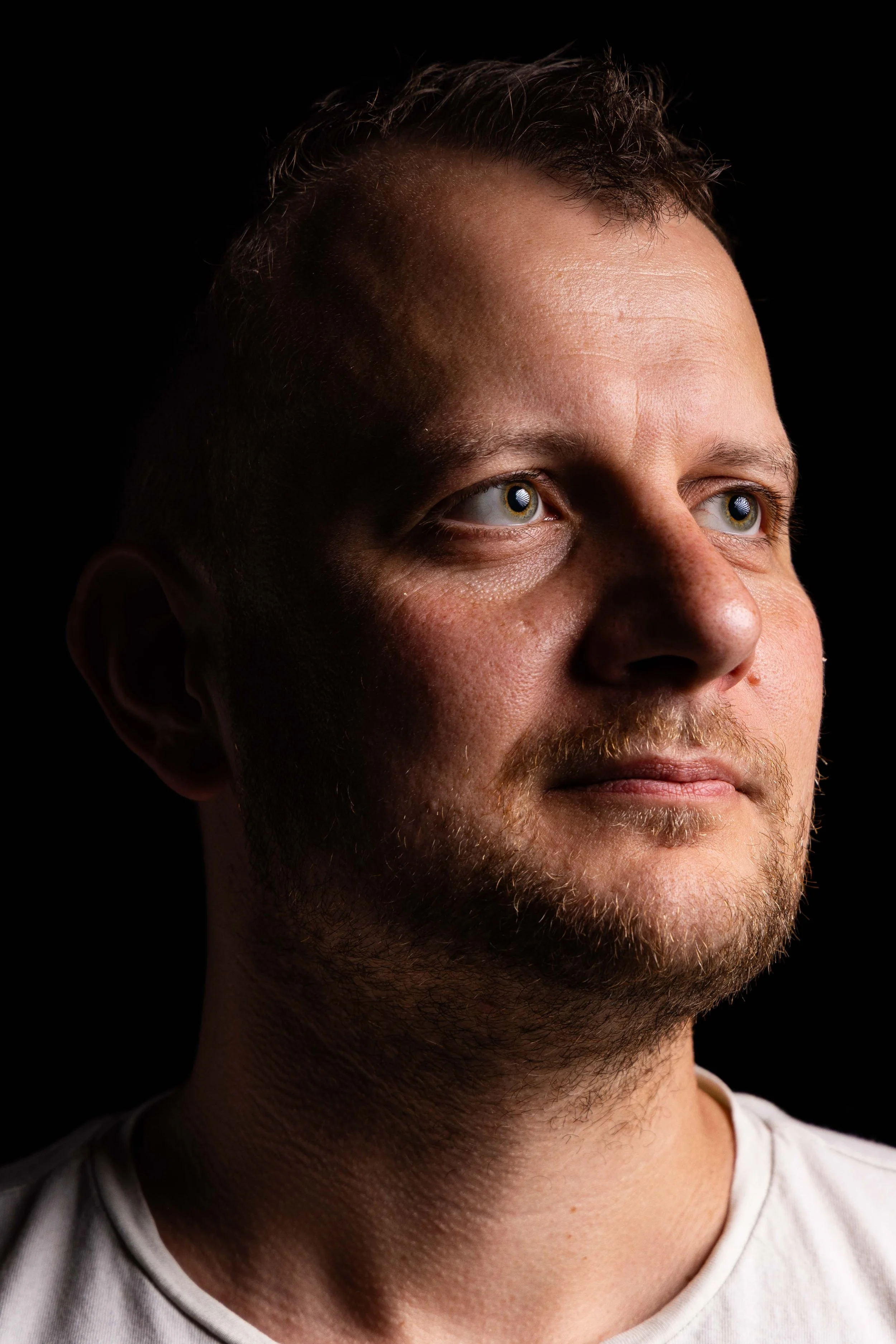 Nahaufnahme eines Mannes mit kurzen Haaren und Bart, Blick nach rechts, dunkler Hintergrund.