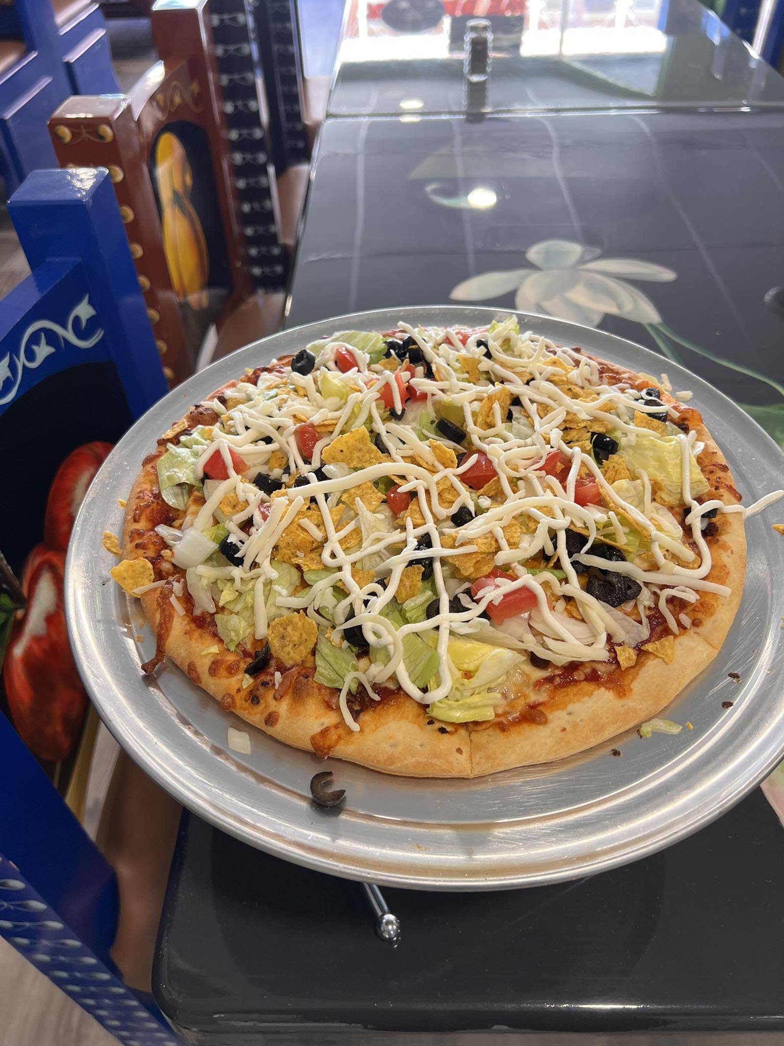 Plate of pizza topped with lettuce, diced tomatoes, black olives, shredded cheese, and tortilla chips on a metal tray.