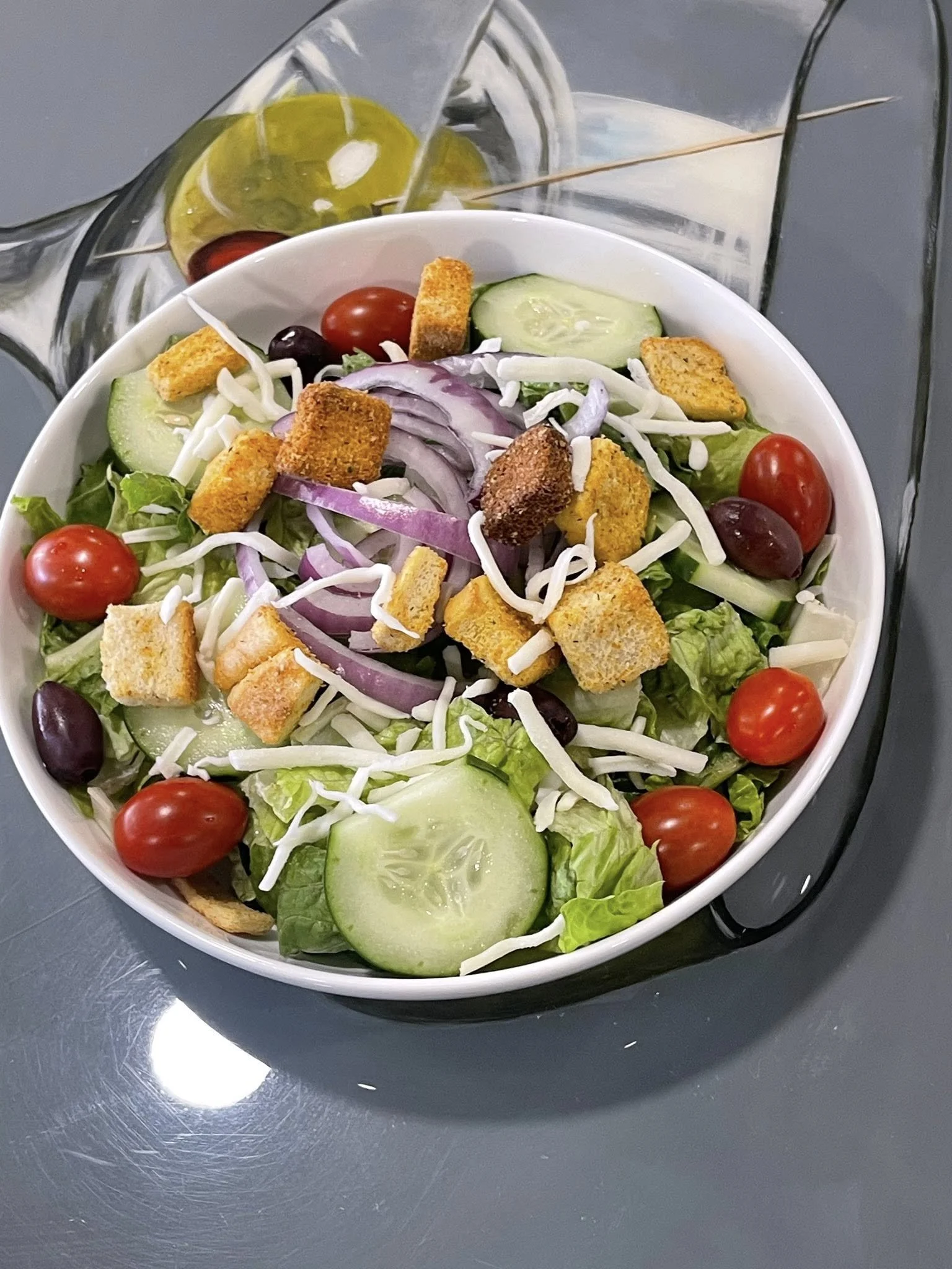 A bowl of fresh salad with cherry tomatoes, cucumber slices, shredded cheese, red onion slices, croutons, and olives, with a glass of water with lemon slices in the background.