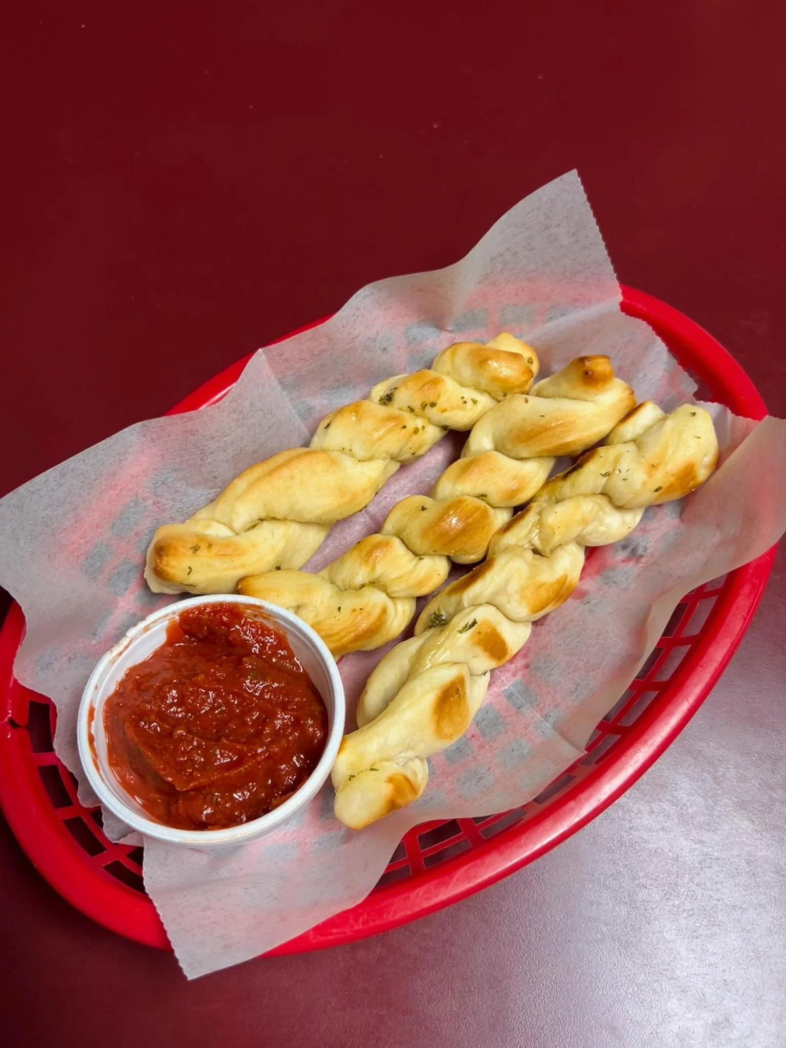 Basket with twisted breadsticks served with marinara sauce in a small cup, on a red tray lined with parchment paper.