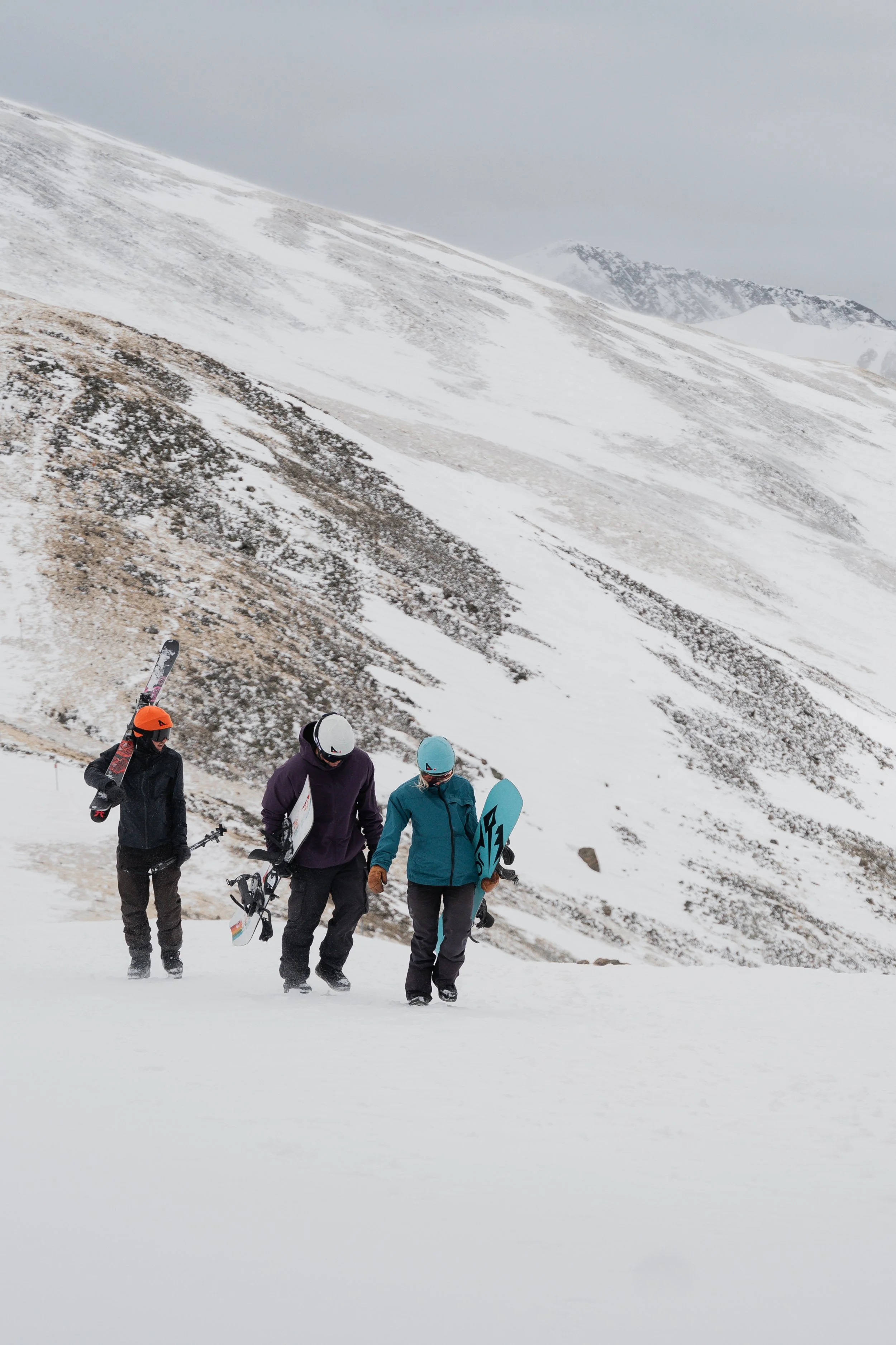 Three snowboarders walking up a snowy mountain slope, carrying snowboards, with a snowy mountain landscape in the background.
