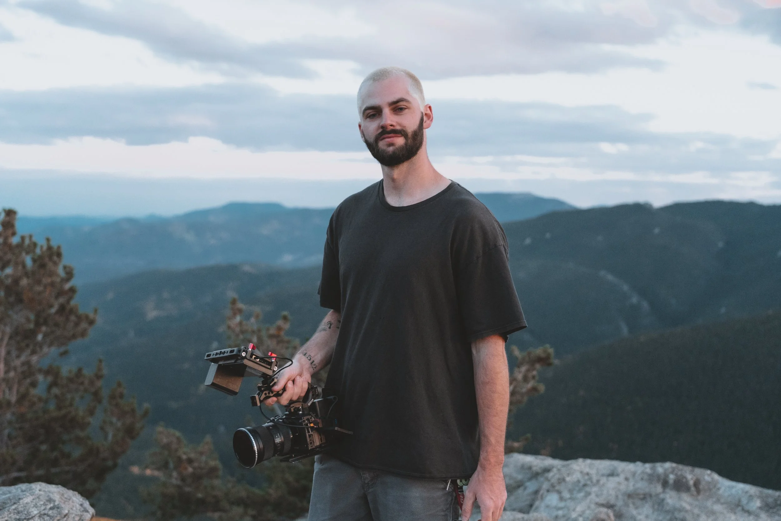 Man standing on rocky ledge holding a professional camera with a mountain landscape in the background.
