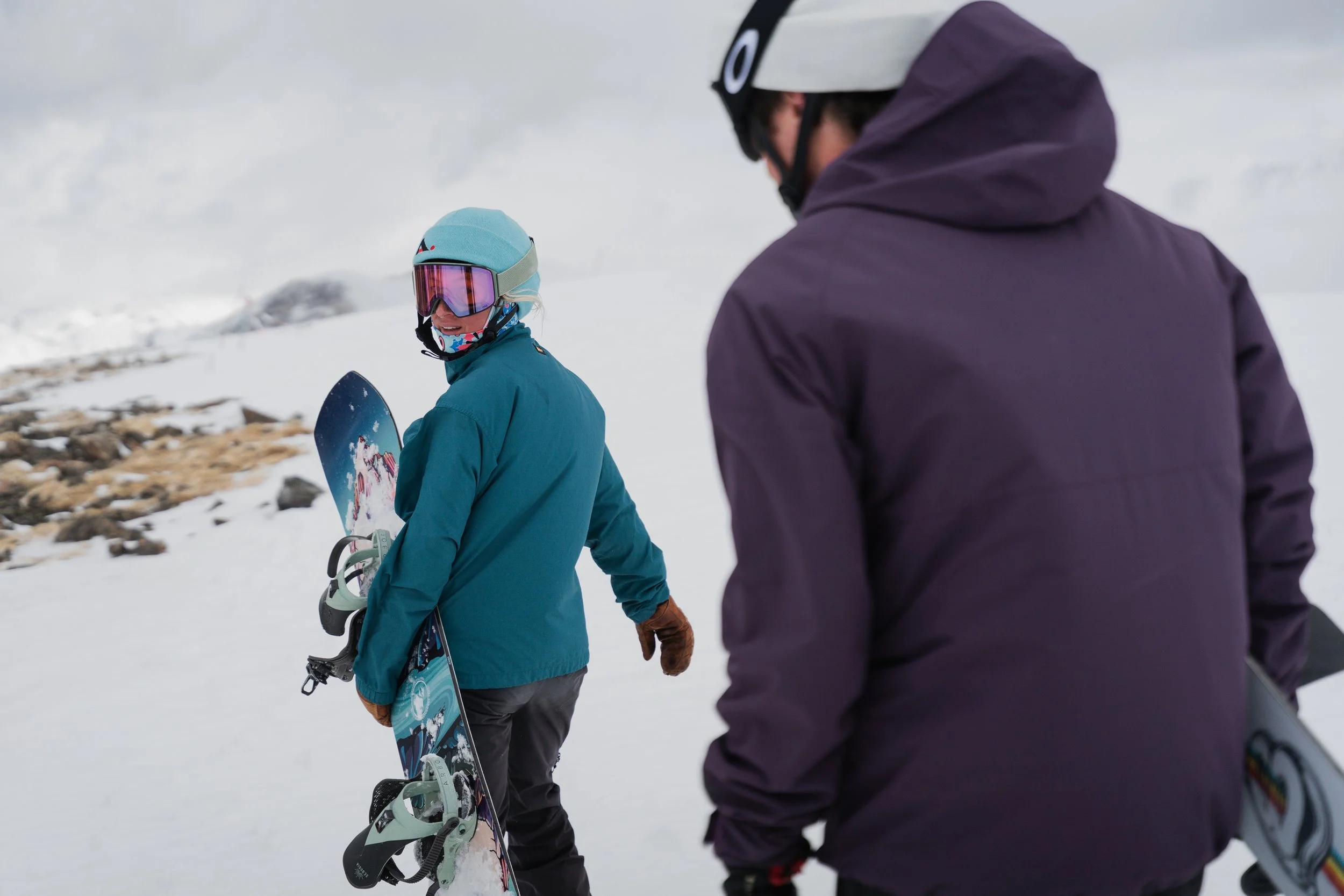 Two people in winter gear, one holding a snowboard, standing on snow in a mountainous area, with one looking over their shoulder and smiling.