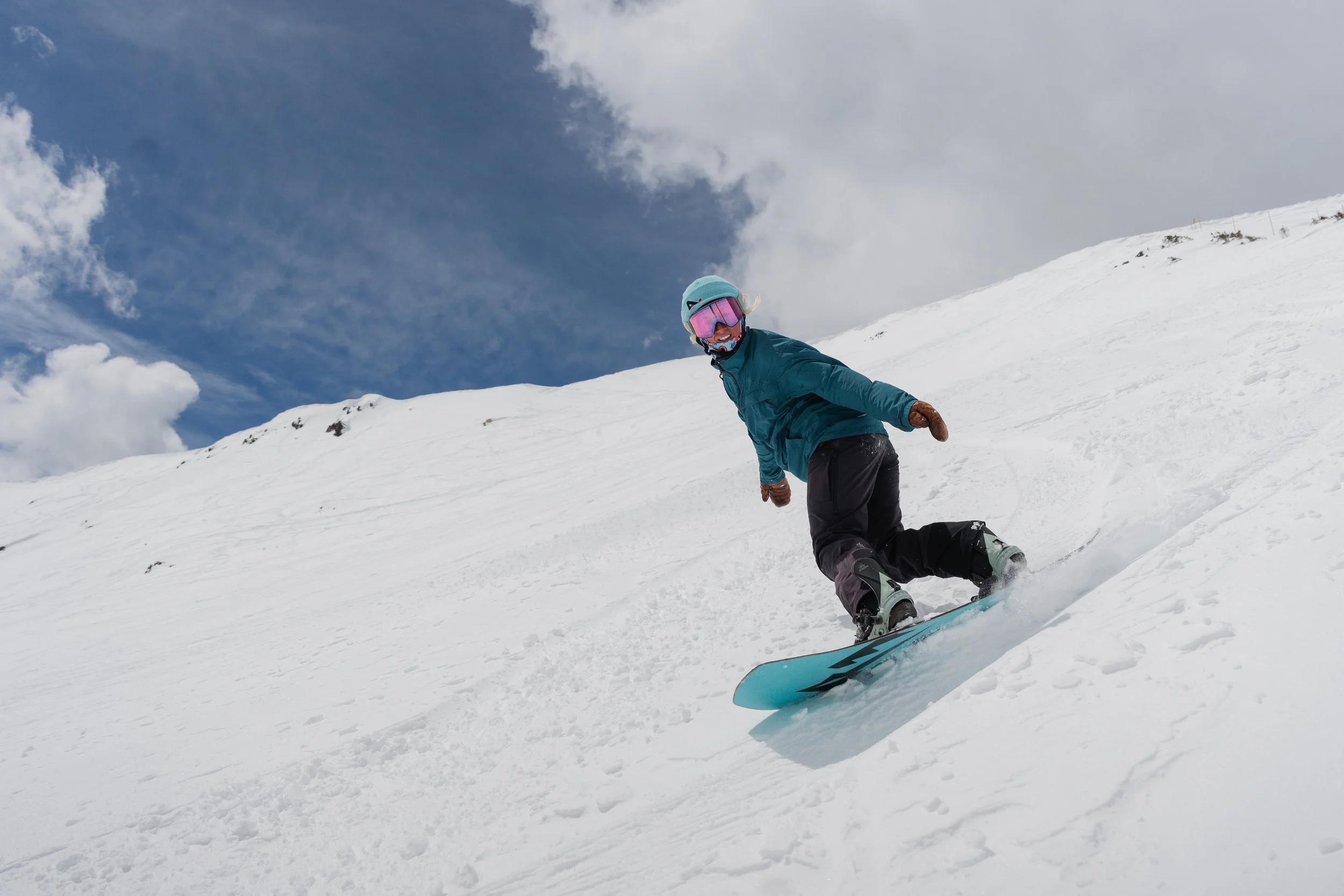 A person snowboarding downhill on a snowy mountain slope, wearing a blue helmet, goggles, teal jacket, black pants, and brown gloves, with a partly cloudy sky in the background.
