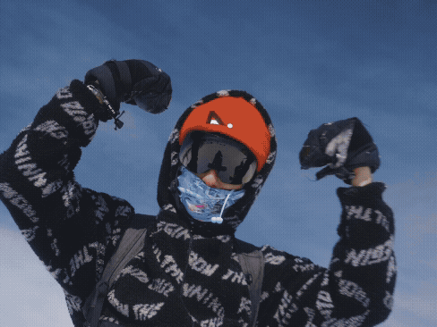Person dressed in winter gear, wearing an orange helmet, goggles, and a mask, raising fists in a victorious pose against a clear blue sky.