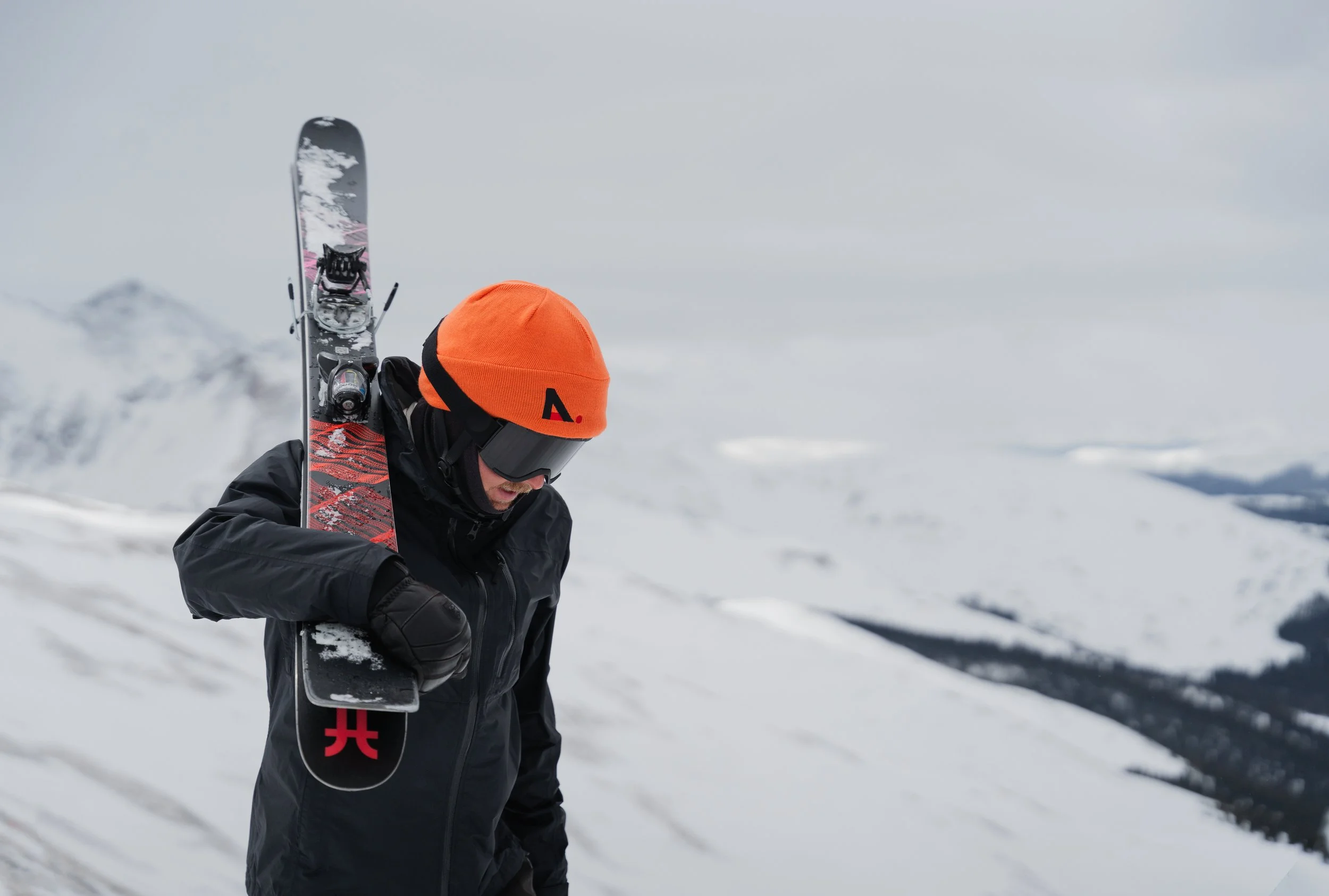 A skier wearing a black jacket, orange beanie, and goggles carries skis over his shoulder while walking through a snowy landscape with mountains in the background.