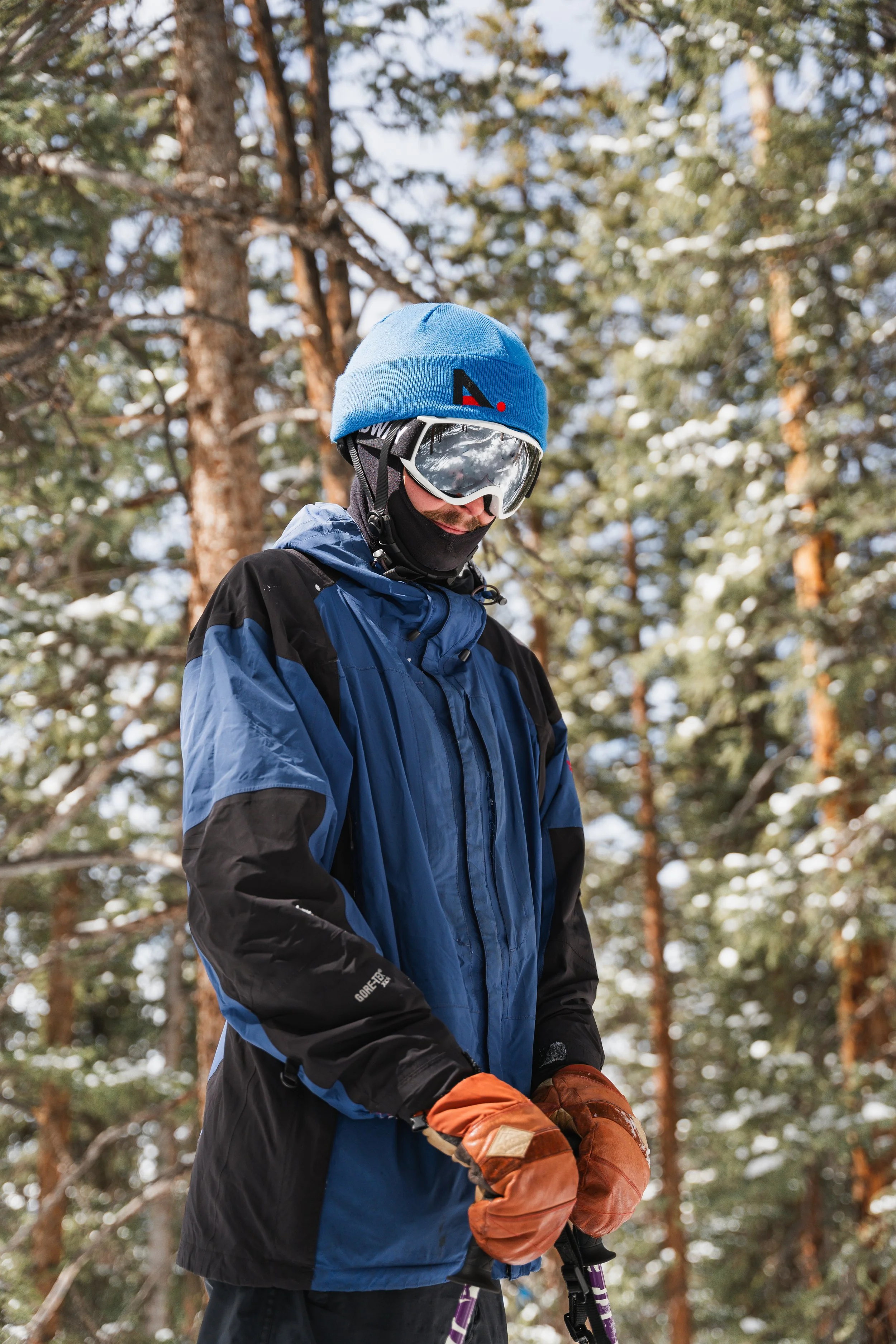 A person dressed in winter gear, including a blue jacket, orange gloves, and a blue beanie, standing outdoors in a snow-covered forest with tall trees.
