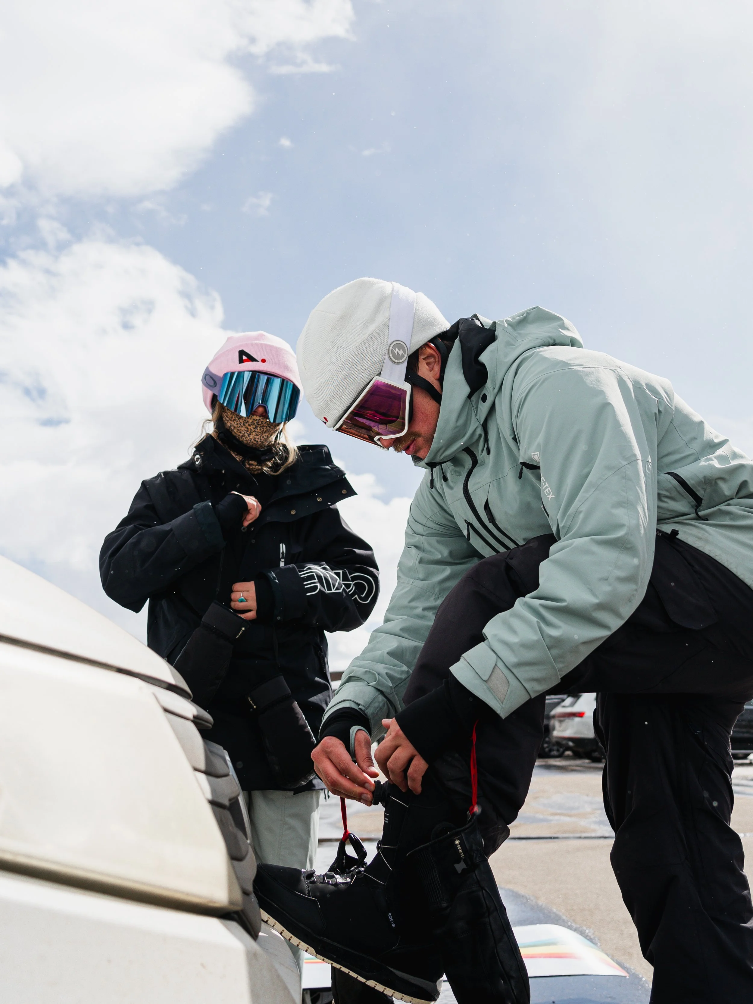 Two people dressed in winter and ski gear, preparing for skiing in a parking lot with cars and a snowy mountain background.