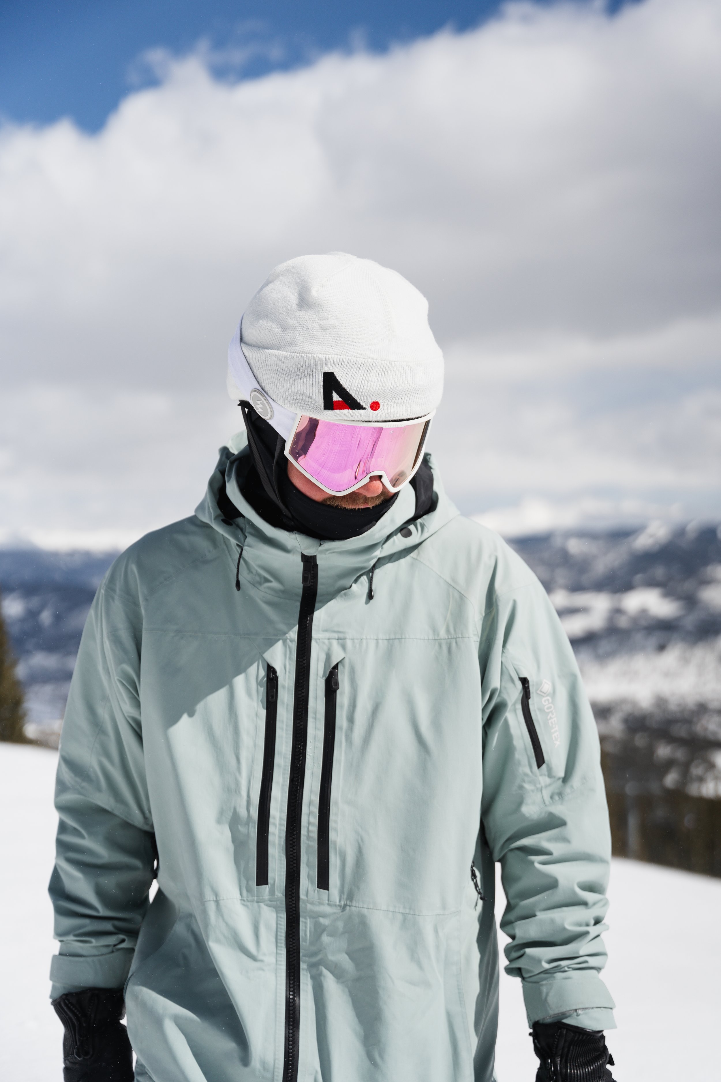 A man dressed in winter ski gear, including a grey jacket, white beanie, and ski goggles, outdoors in a snowy mountain landscape with cloudy sky.