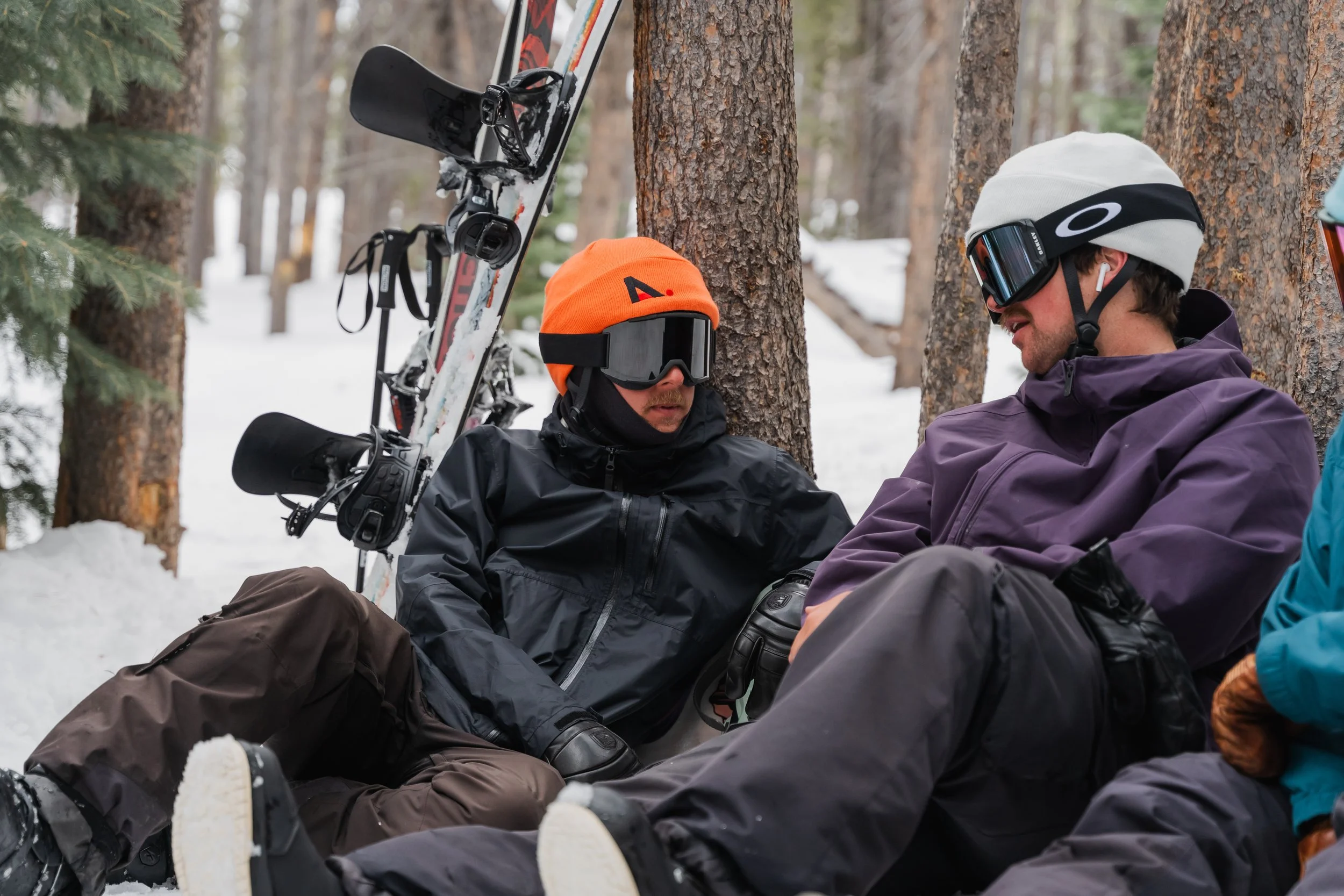 Two skiers resting on the snow in a forest, sitting against trees, with a pair of skis and ski poles nearby.