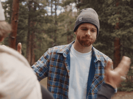 A young man with a beard wearing a gray beanie, plaid shirt, and white T-shirt outdoors in a forest, gesturing with his hands.