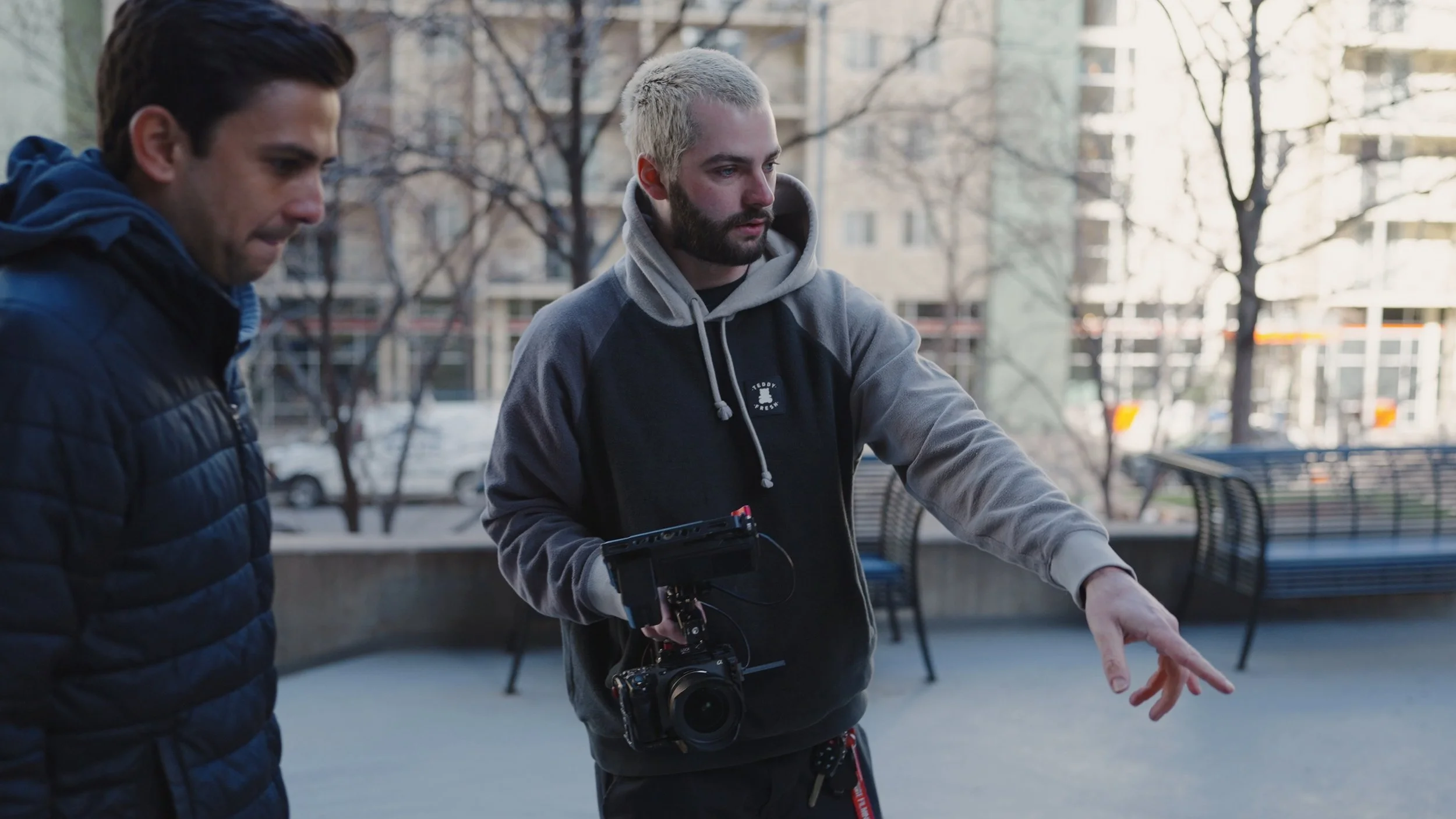 Two men engaged in a discussion outdoors in an urban park setting, one holding a camera and a monitor, with benches and high-rise buildings in the background during daytime.