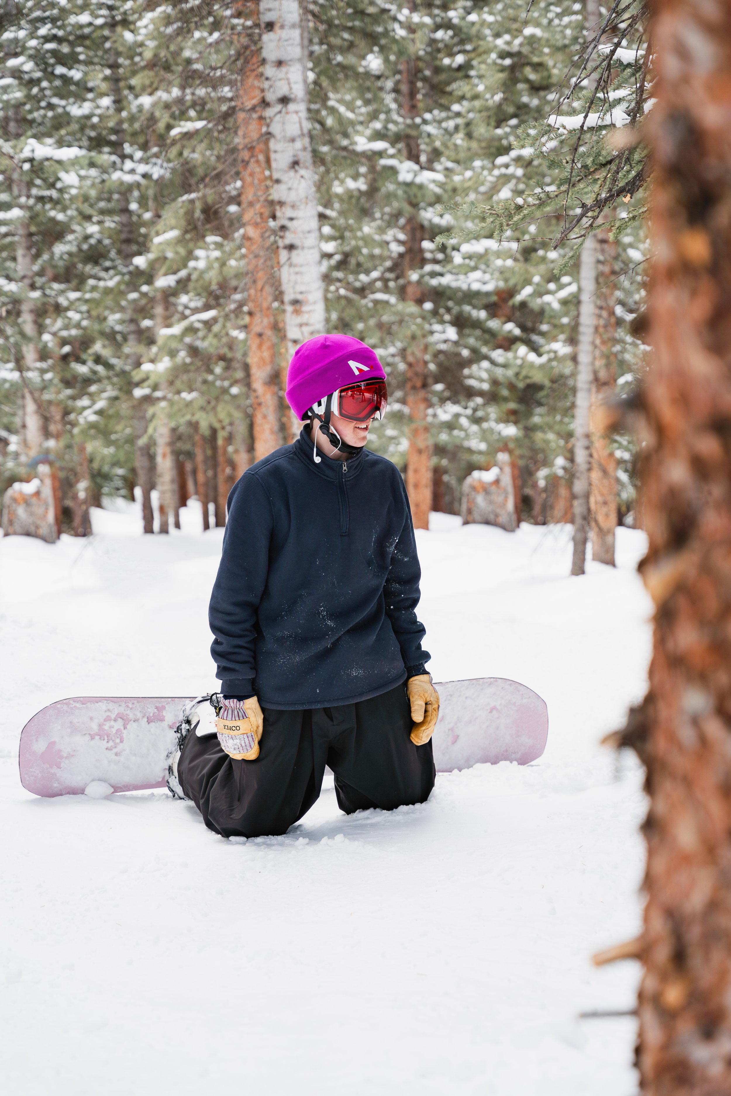A person kneeling in the snow in a forest, wearing a purple beanie, goggles, black winter jacket, and holding a snowboard.