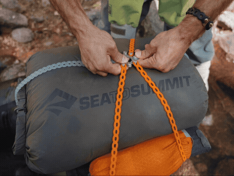 Person fastening a zipper on a gray SEATOSUMIT sleeping bag with orange straps, outdoors on rocky terrain.