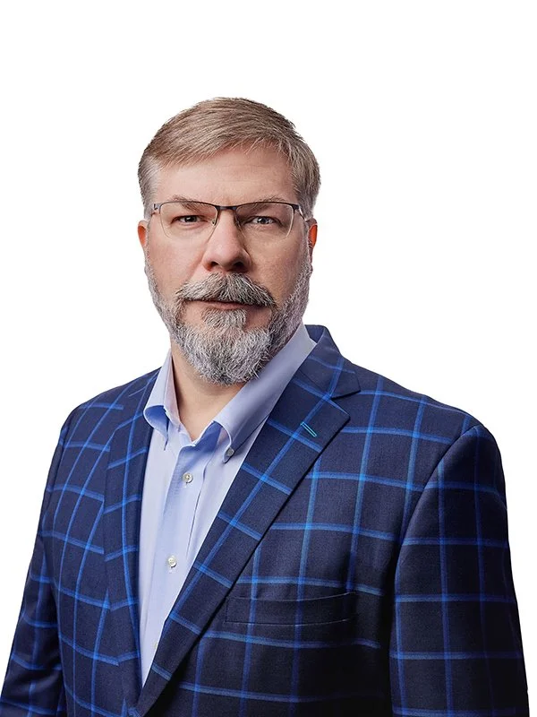 A middle-aged man with glasses, a gray beard, and mustache wearing a blue checkered blazer and light blue shirt against a white background.