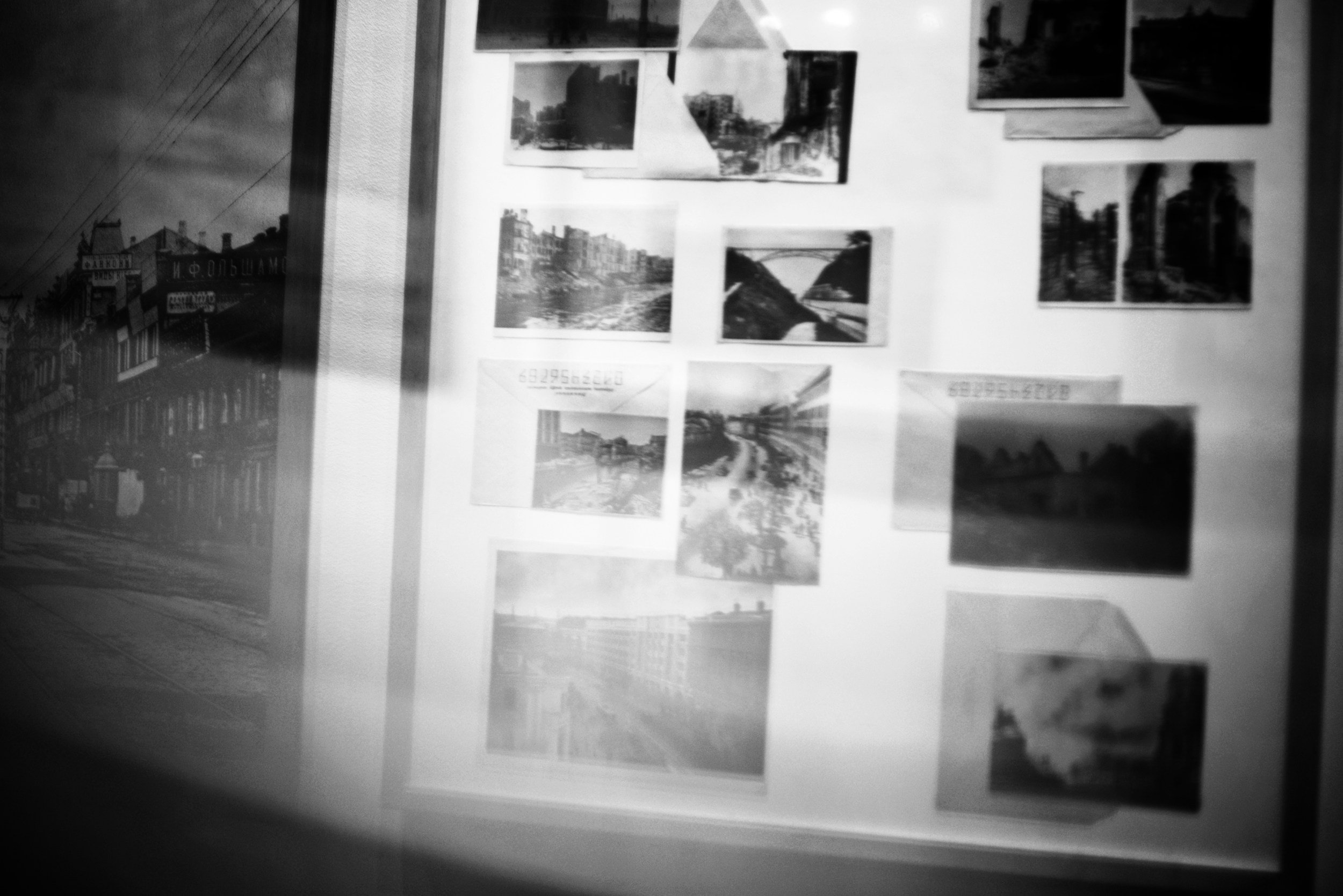 Black and white photo of a wall displaying various framed photographs of cityscapes and landscapes, with a window or mirror reflection on the left showing a building with sign and power lines.