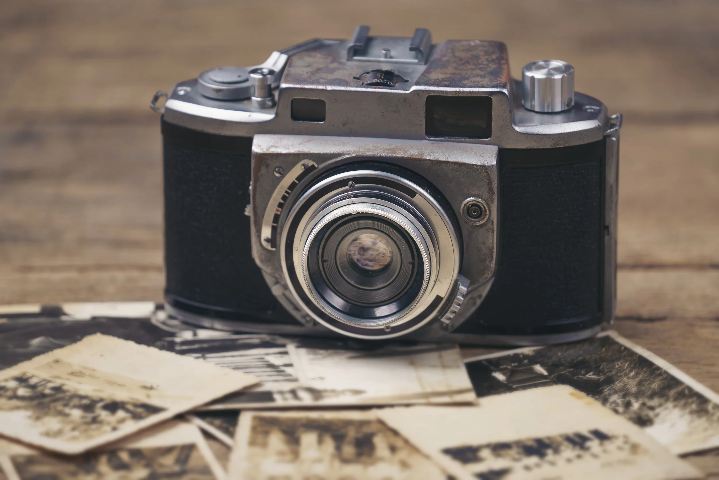 A vintage camera with a rusted metal body sits on a wooden surface covered with black and white printed photographs or papers.