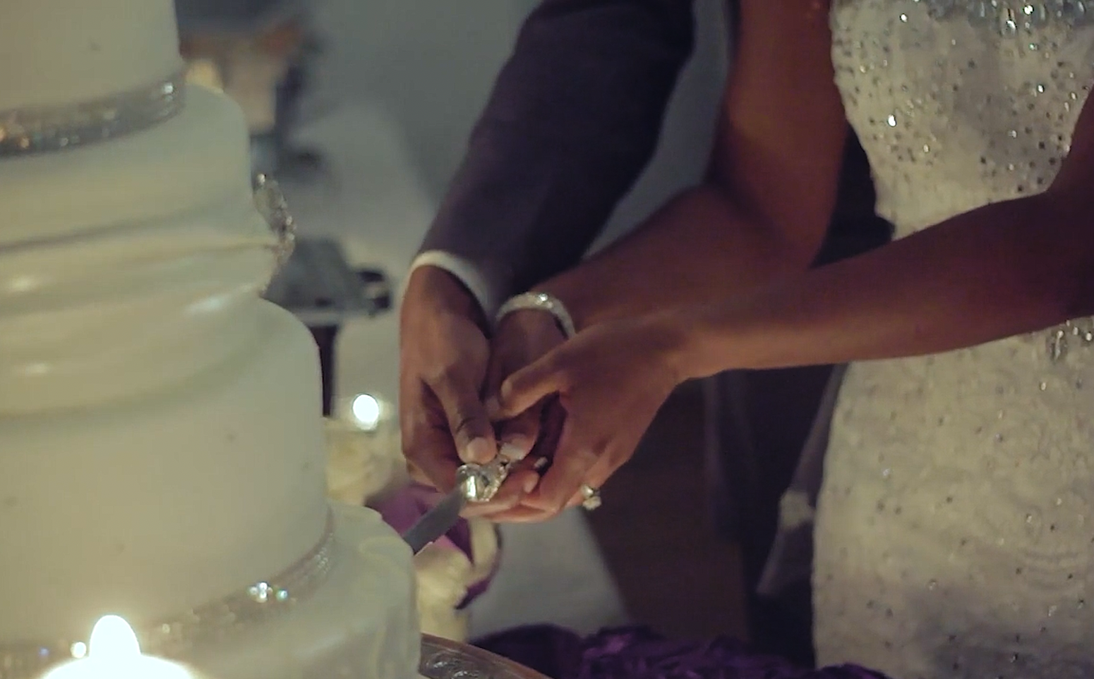 Hands of a couple cutting a wedding cake together, with wedding attire and jewelry visible.