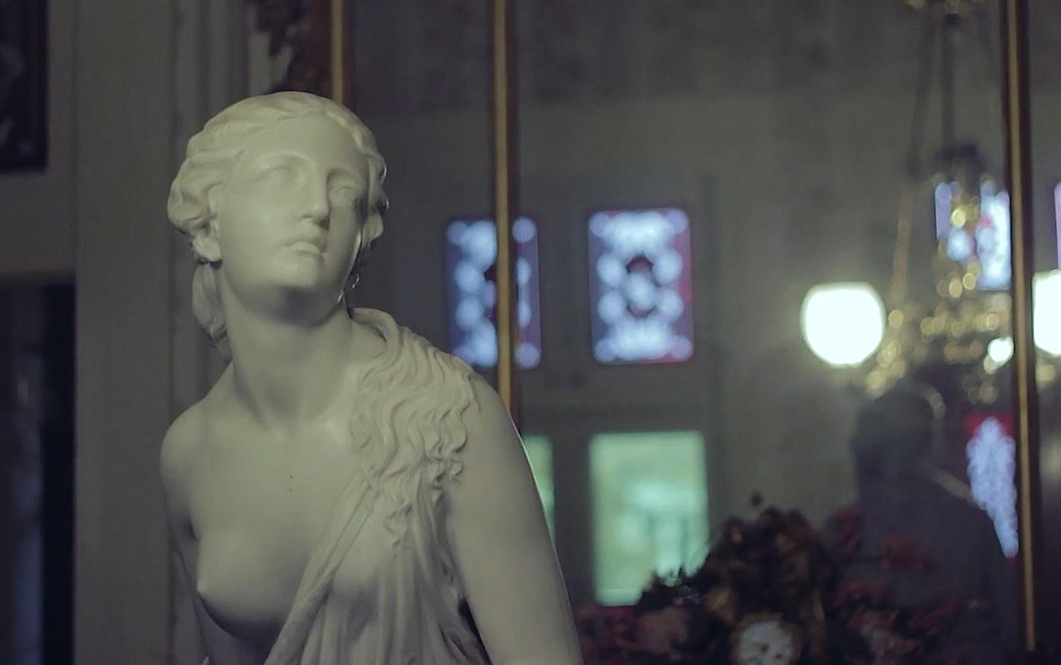 A white marble bust sculpture of a woman with long flowing hair, displayed behind a glass or mirror, with a blurred interior background including windows and decorative lights.