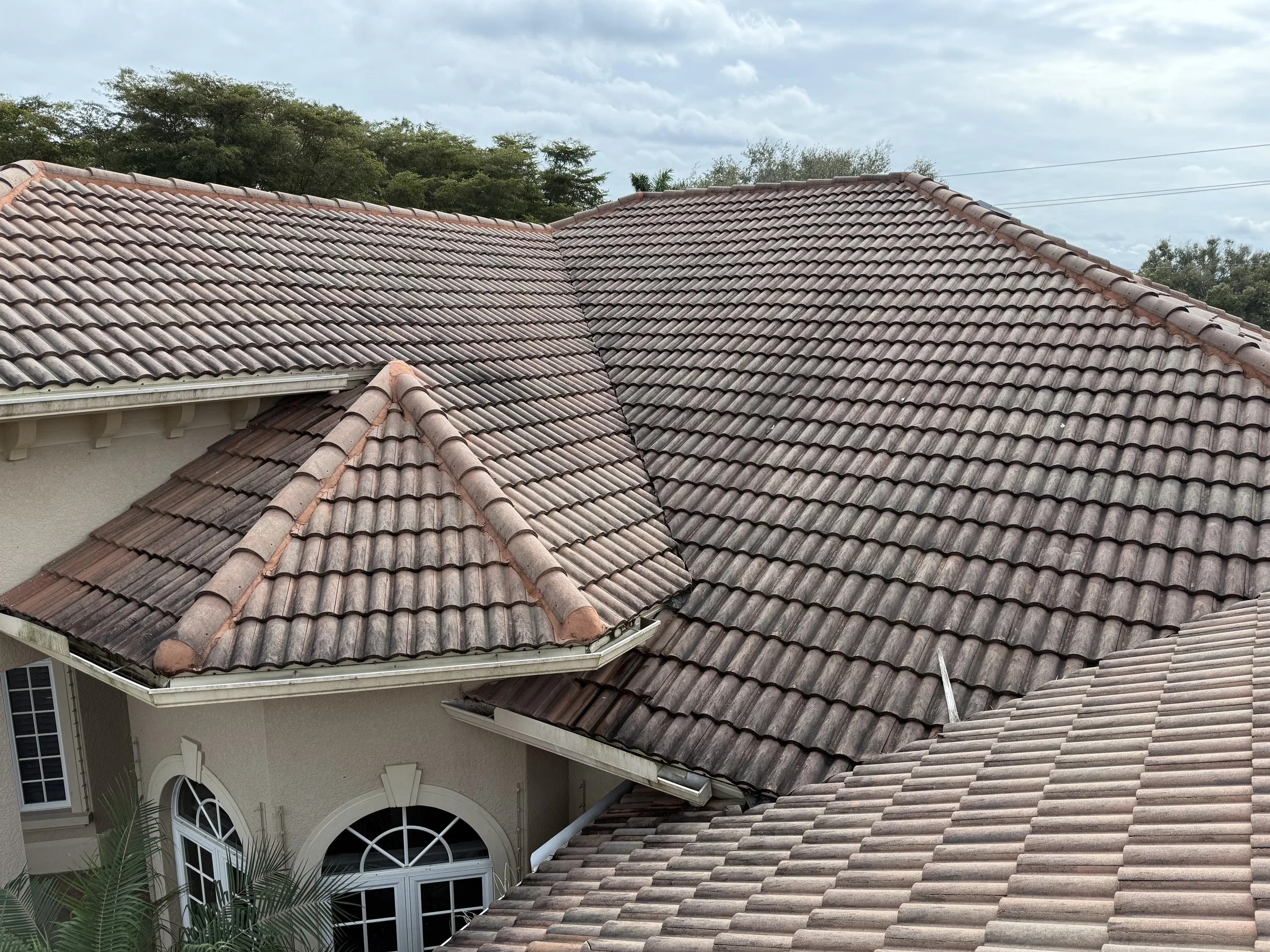 View of multiple overlapping roof sections covered with reddish-brown tiles on a house, with trees and a cloudy sky in the background.