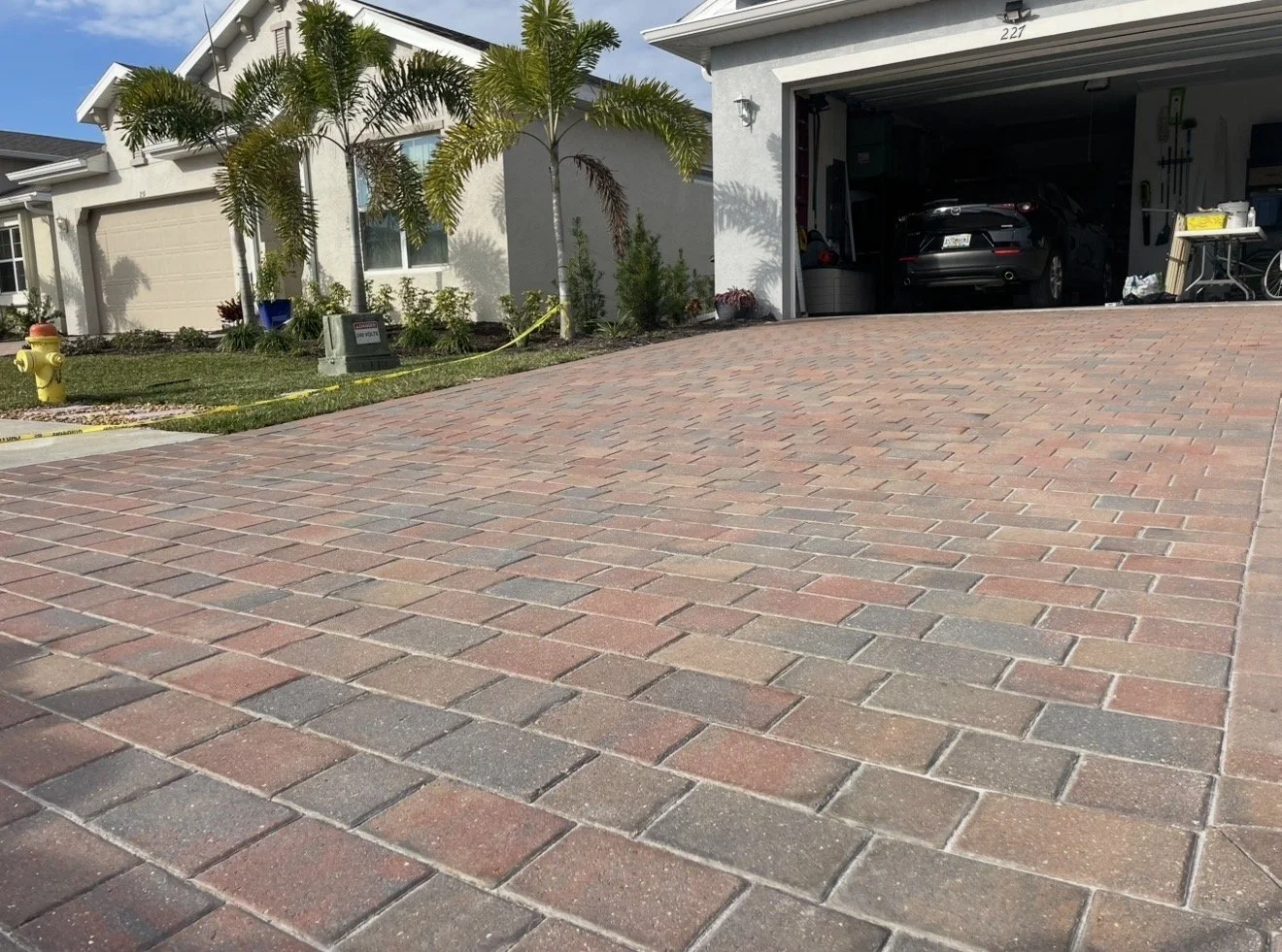 View of a residential driveway paved with interlocking bricks, leading to a garage with a car parked inside. The house has a white exterior, and there are palm trees and greenery in the yard on the left. There is a yellow fire hydrant and a utility b