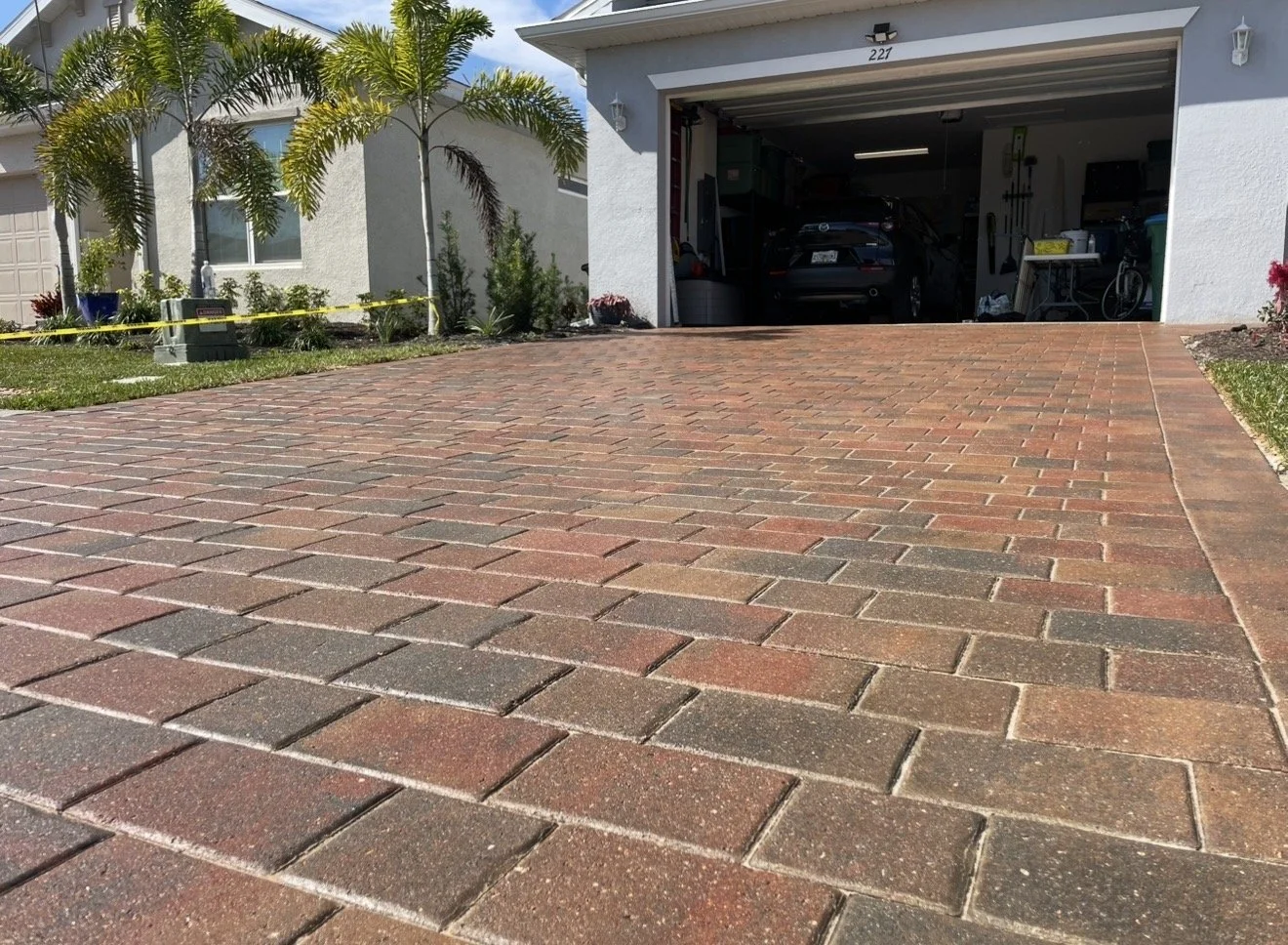View of a residential driveway leading to an open garage with a black car inside, white house exterior, palm trees, and plants in the yard.