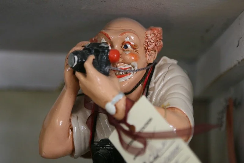 A humorous ceramic figurine of a man with a clown nose and white face paint holding a camera, appearing to take a photo with a smiling expression.