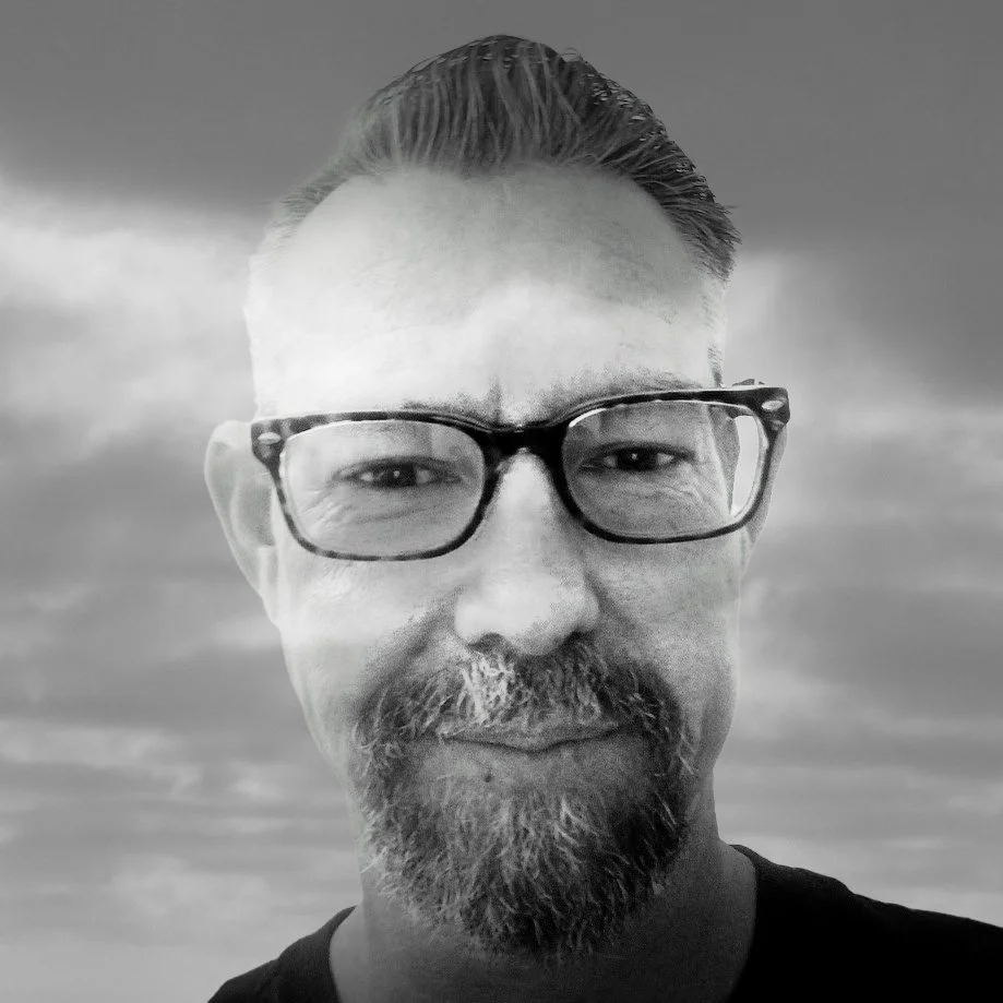 A black and white close-up photo of a man with glasses, a beard, and short hair, smiling slightly against a cloudy sky.