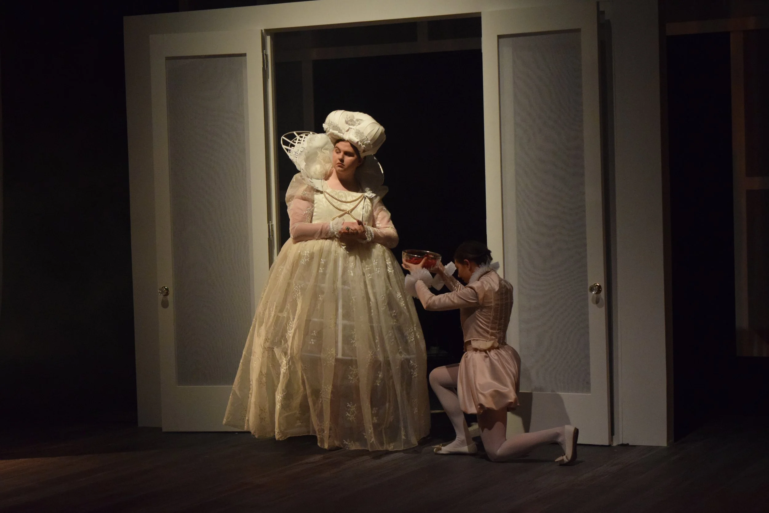 Two women in elaborate period costumes, one standing and one kneeling, with a dark background and open doors behind them.