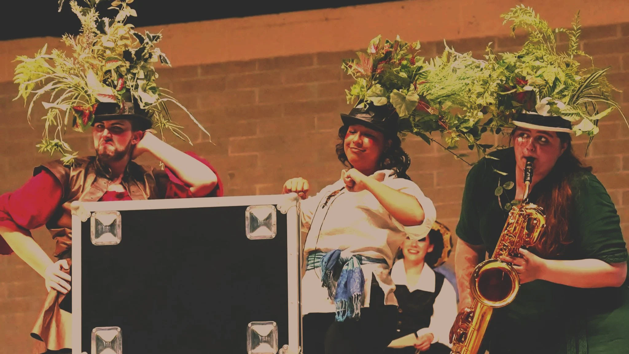 Three performers with headpieces made of greenery and leaves, posing on stage. The first on the left has a large, elaborate headpiece and a serious expression. The middle performer is smiling confidently, with a smaller headpiece and a wrapped scarf. The third on the right is playing a saxophone, with a headpiece adorned with leaves and a focused expression. There is a woman in the background smiling, dressed in a black and white costume.