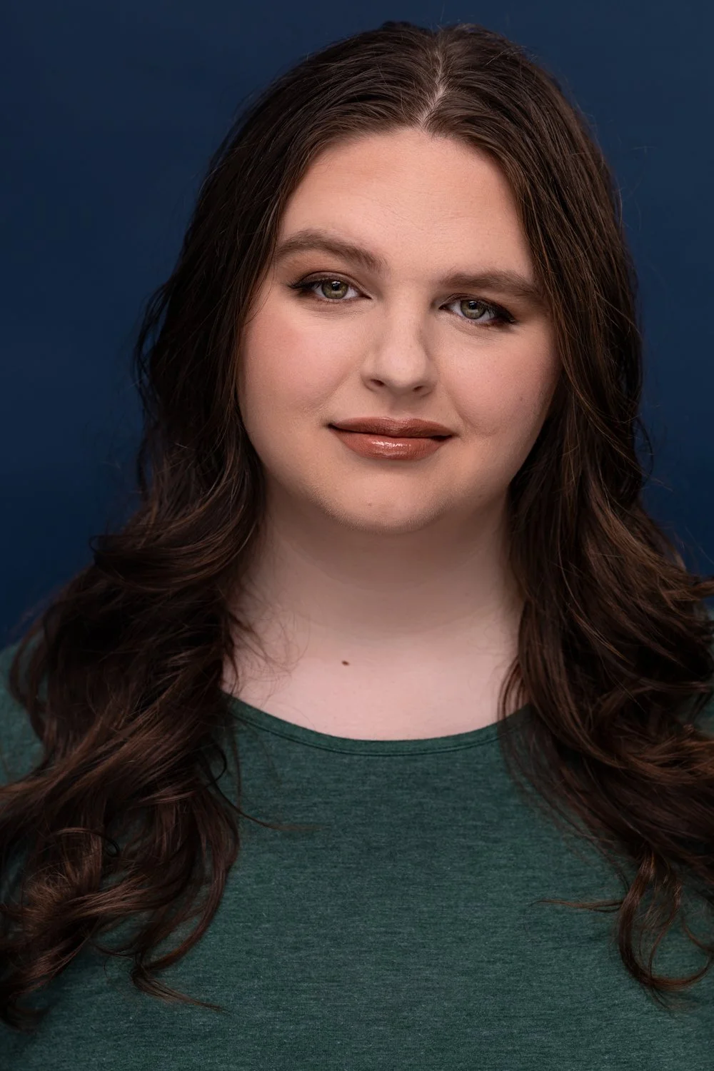 A woman with long, wavy brown hair, light makeup, and a shiny, natural lip color, wearing a dark green top, smiling softly against a dark blue background.