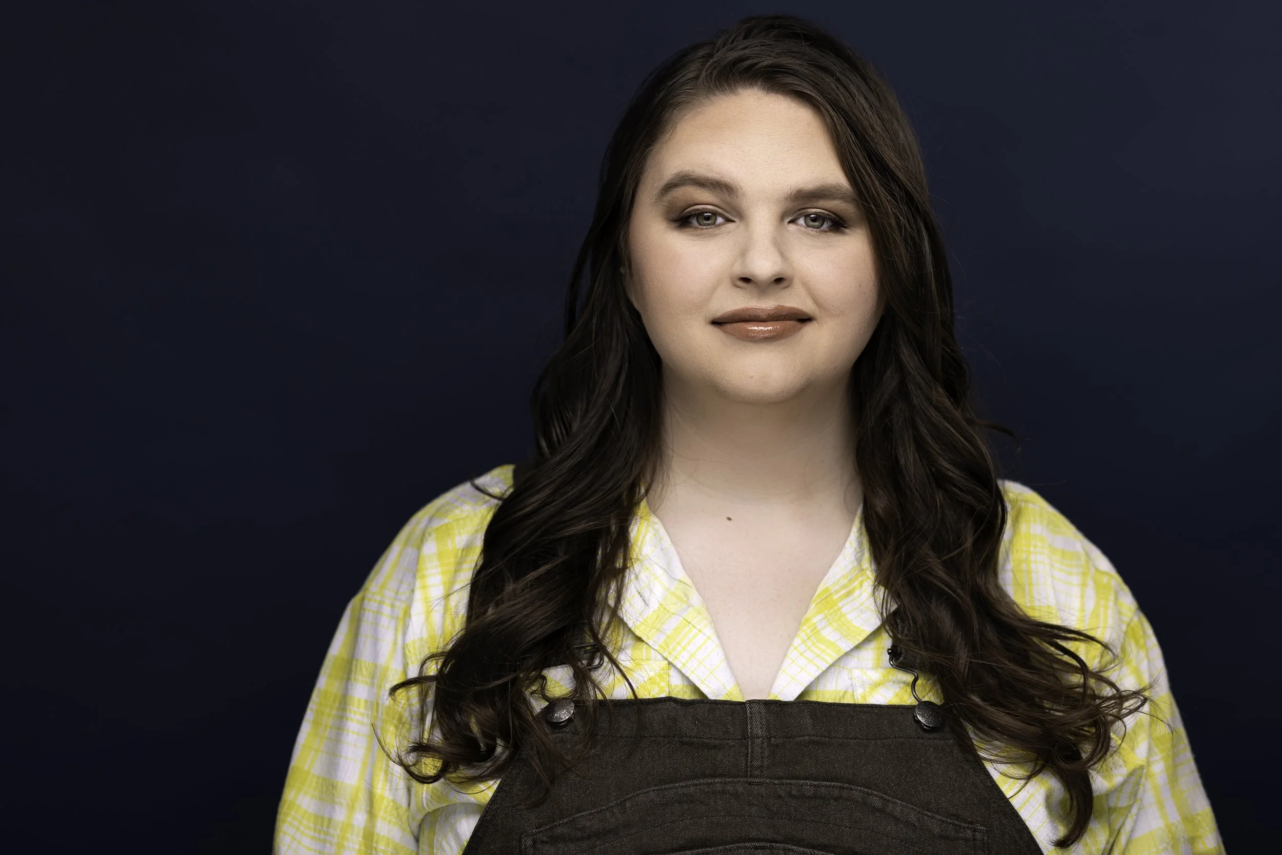 Portrait of a woman with long, wavy brown hair wearing a yellow checkered shirt and brown overalls, standing against a dark background.