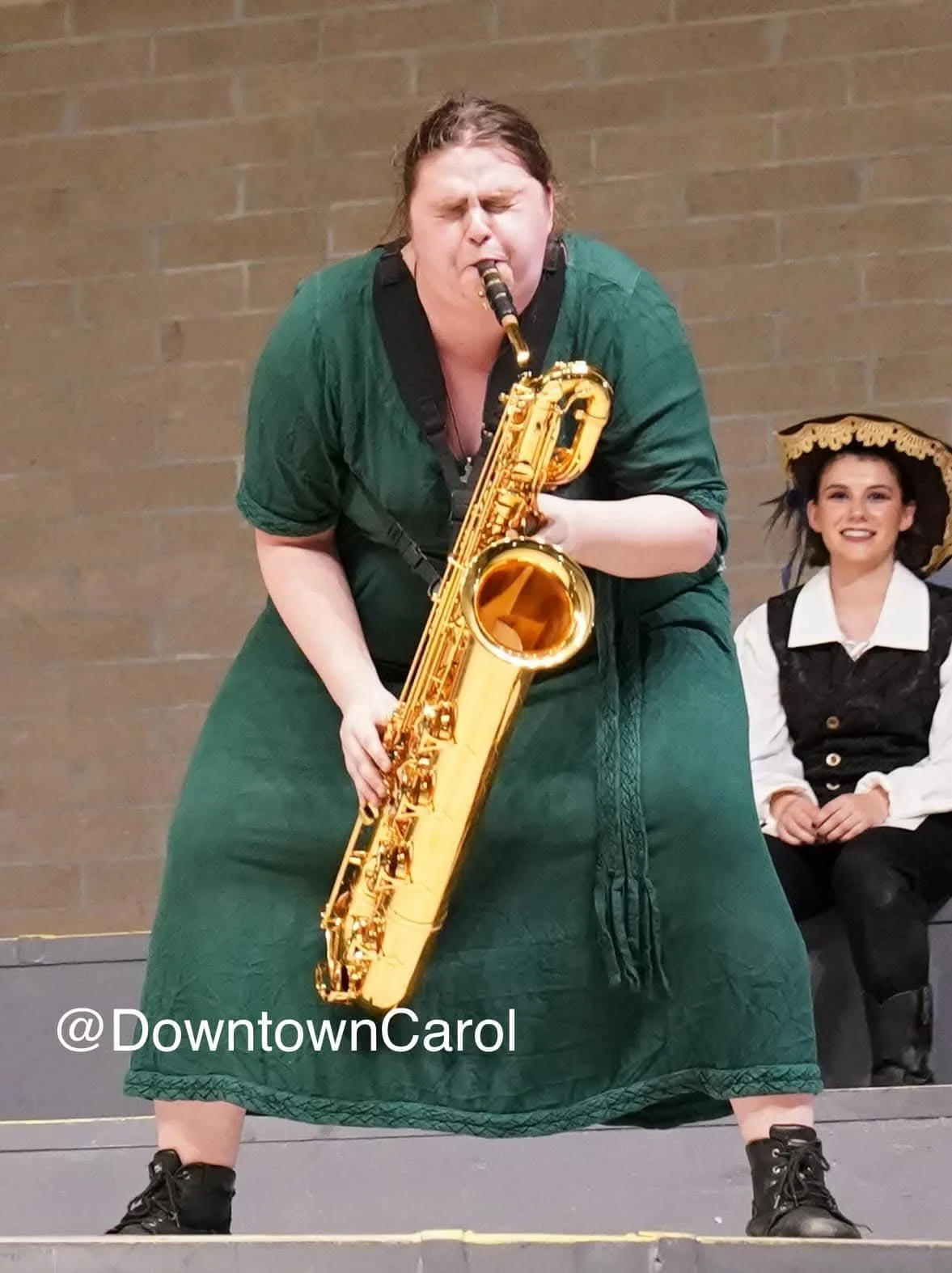 A woman in a green dress passionately playing a saxophone on stage, with a woman in a white blouse, black vest, and large hat sitting behind her and smiling.