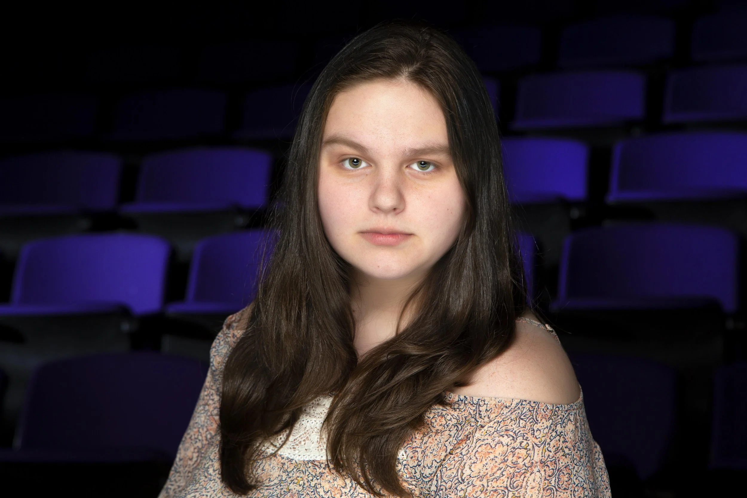 A young woman with long brown hair and fair skin looking directly at the camera, sitting in an auditorium with dark purple seats in the background.