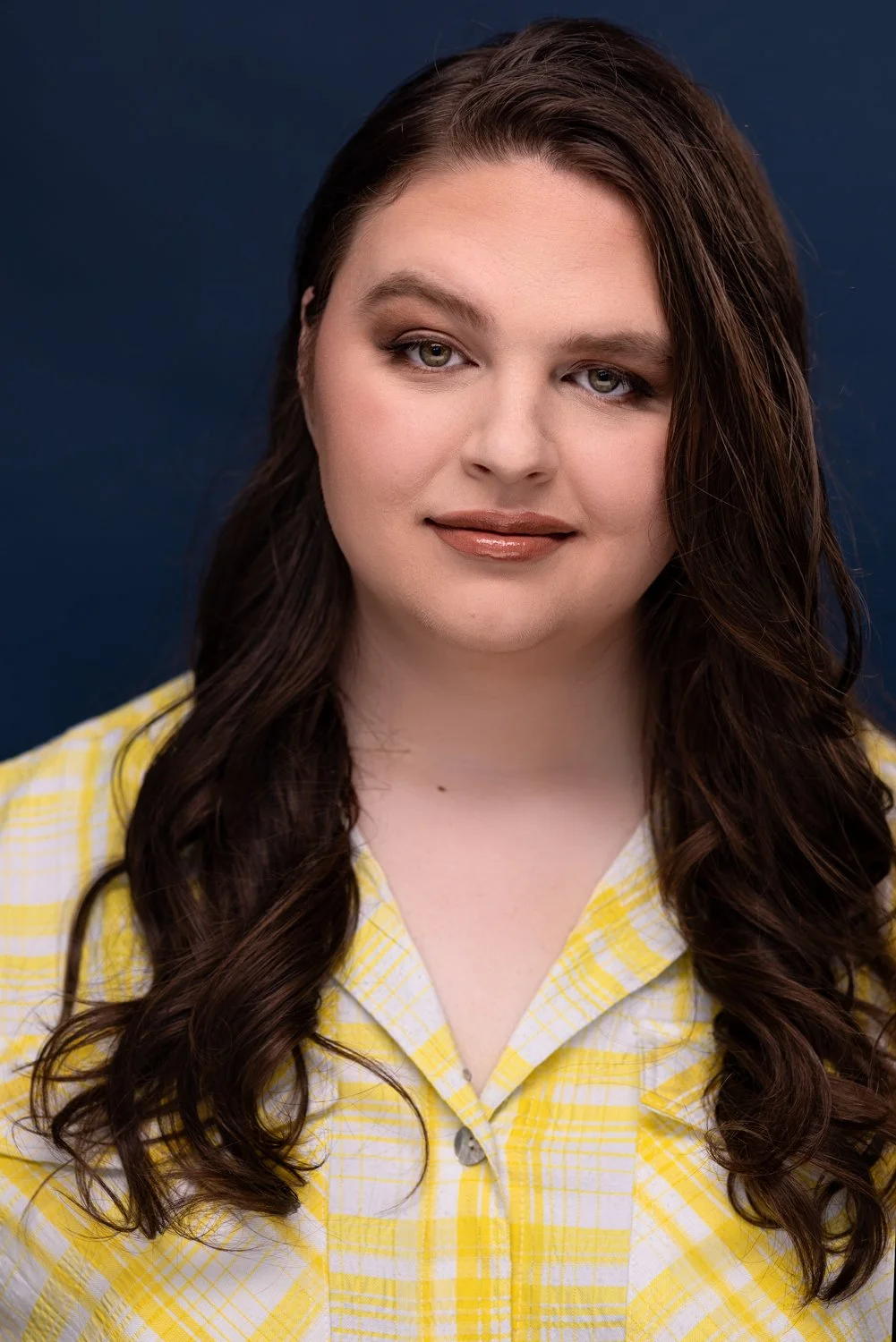 A woman with long, wavy brown hair and fair skin, wearing a yellow plaid shirt, against a dark blue background.