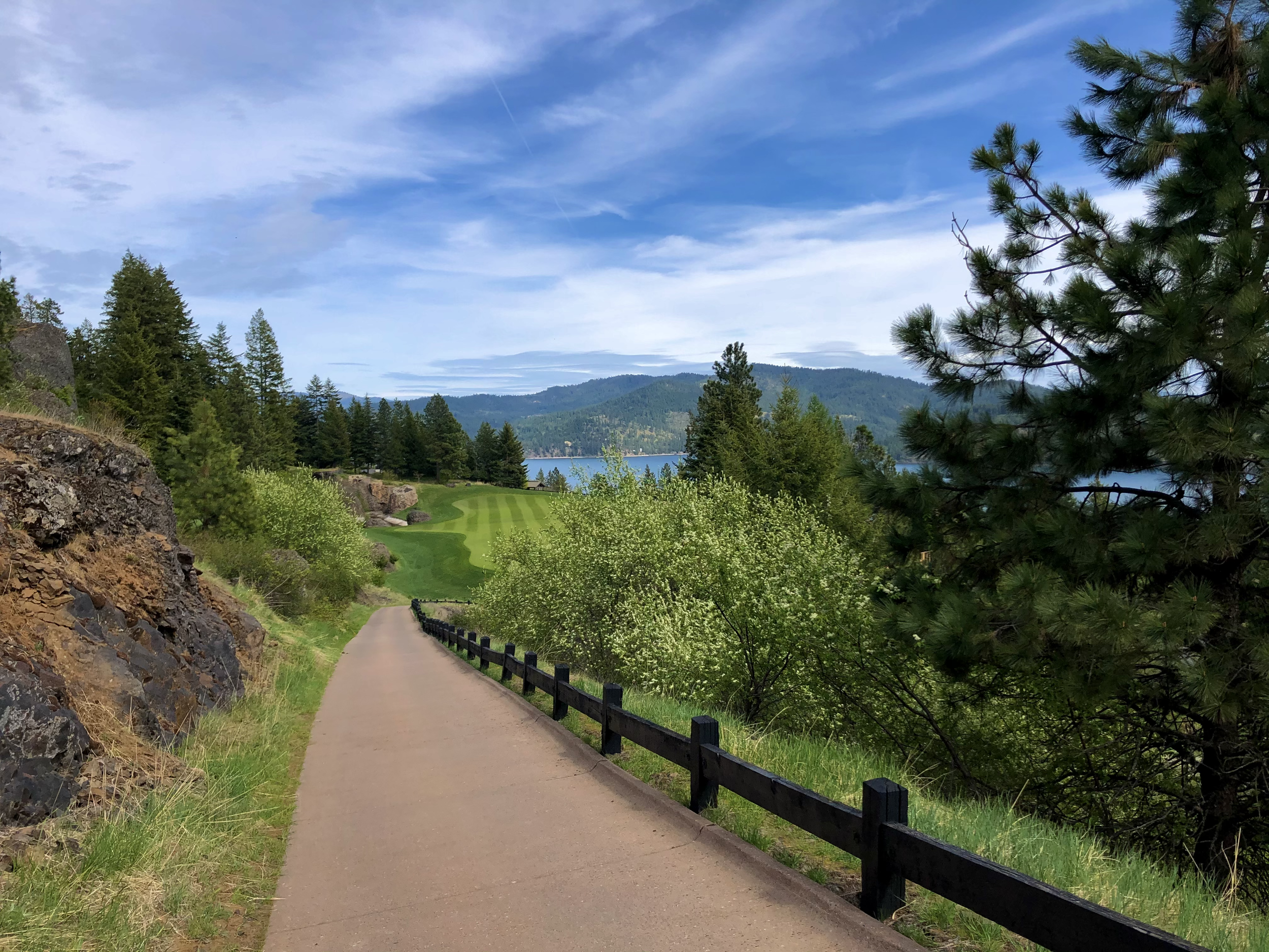 A paved pathway running along a hillside with a wooden fence on the right, surrounded by trees and greenery, leading toward a golf course and a lake with mountains in the distance under a partly cloudy sky.