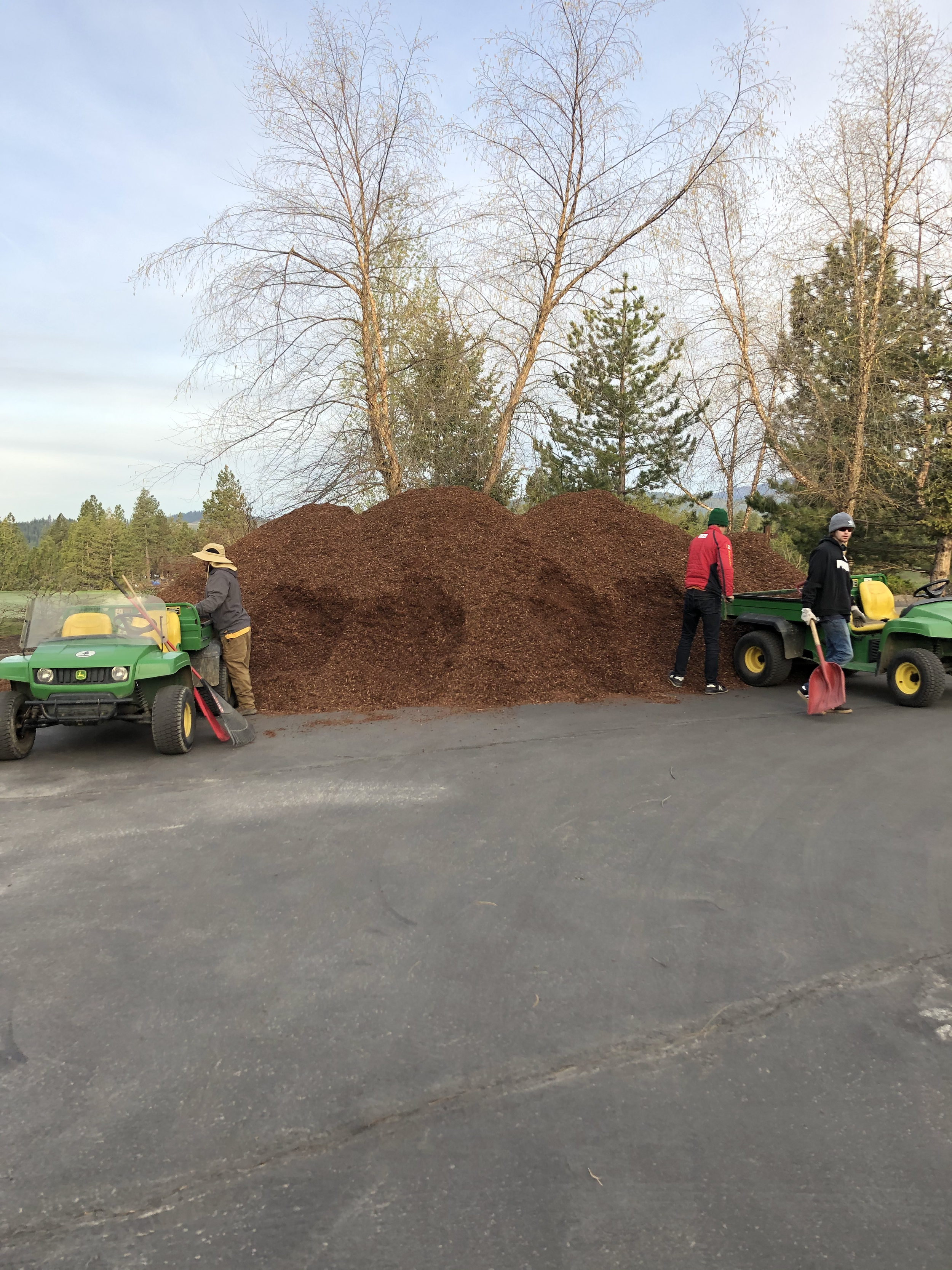 Three people working outdoors with two green utility vehicles parked on a paved surface next to large piles of mulch, with leafless trees in the background.