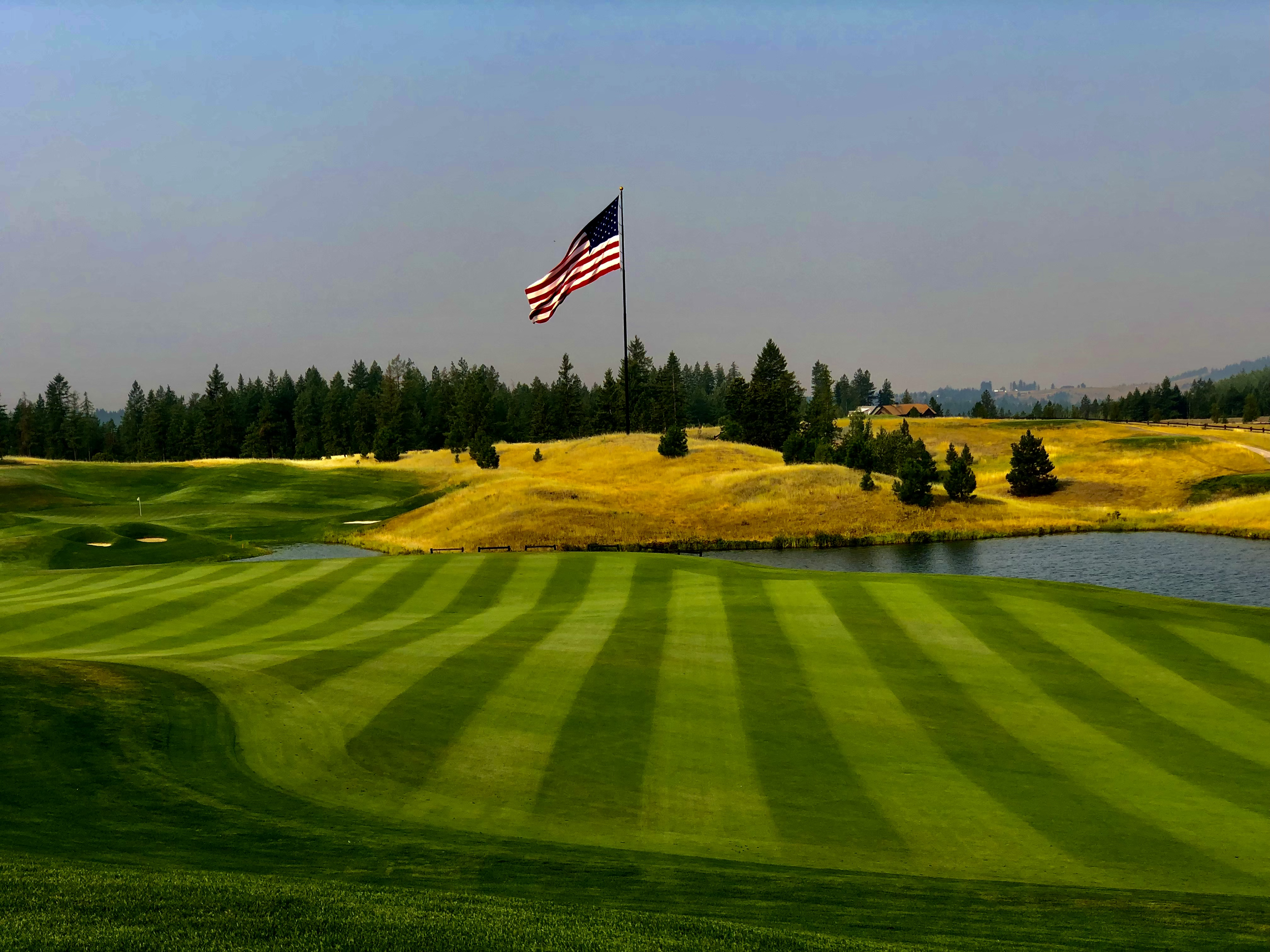 A golf course with a flag on a green, rolling hills in the background, trees, a lake, and an American flag flying.