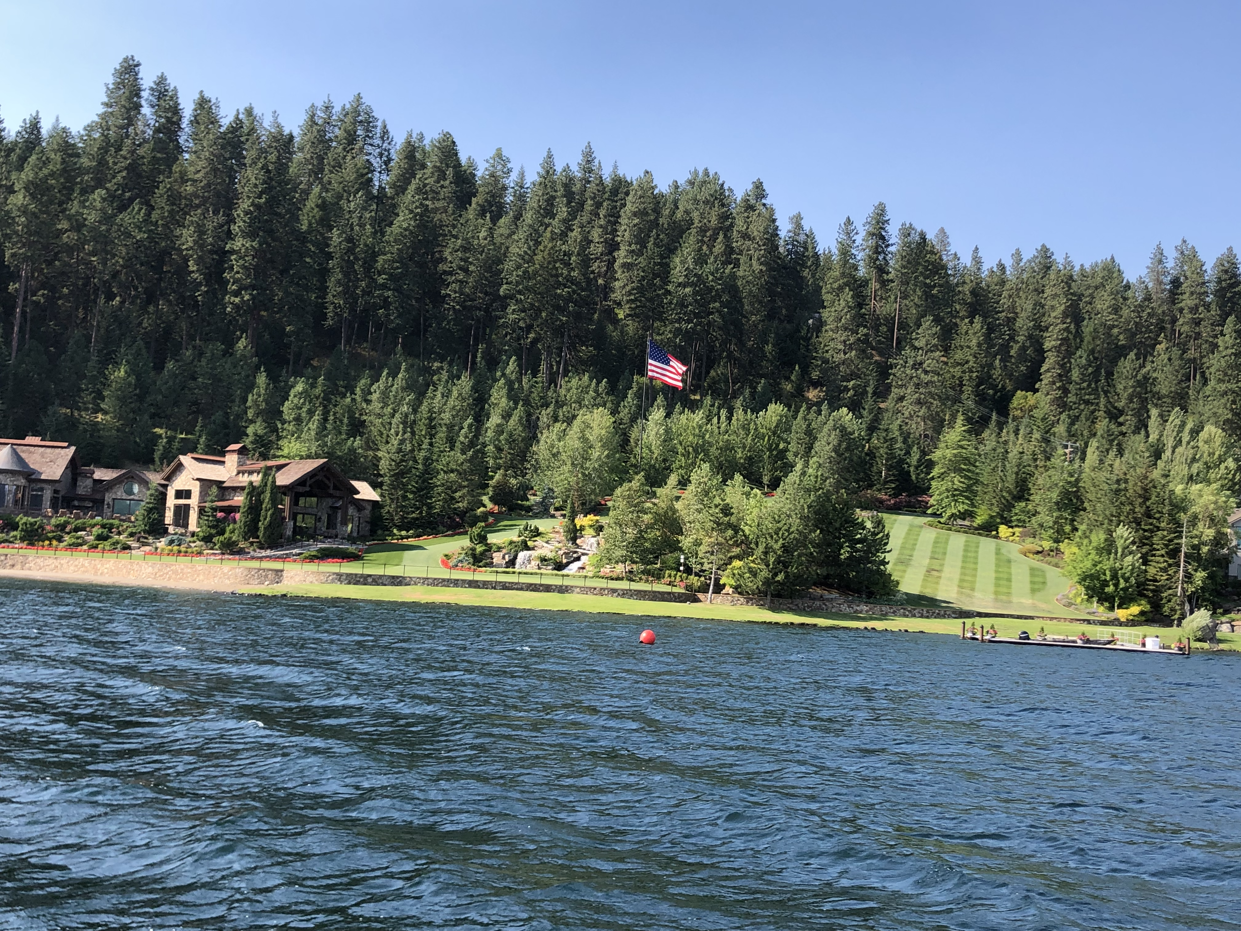 Waterfront view of a property with houses, a well-manicured lawn, trees, and an American flag on a hill, with a lake in the foreground under a clear blue sky.